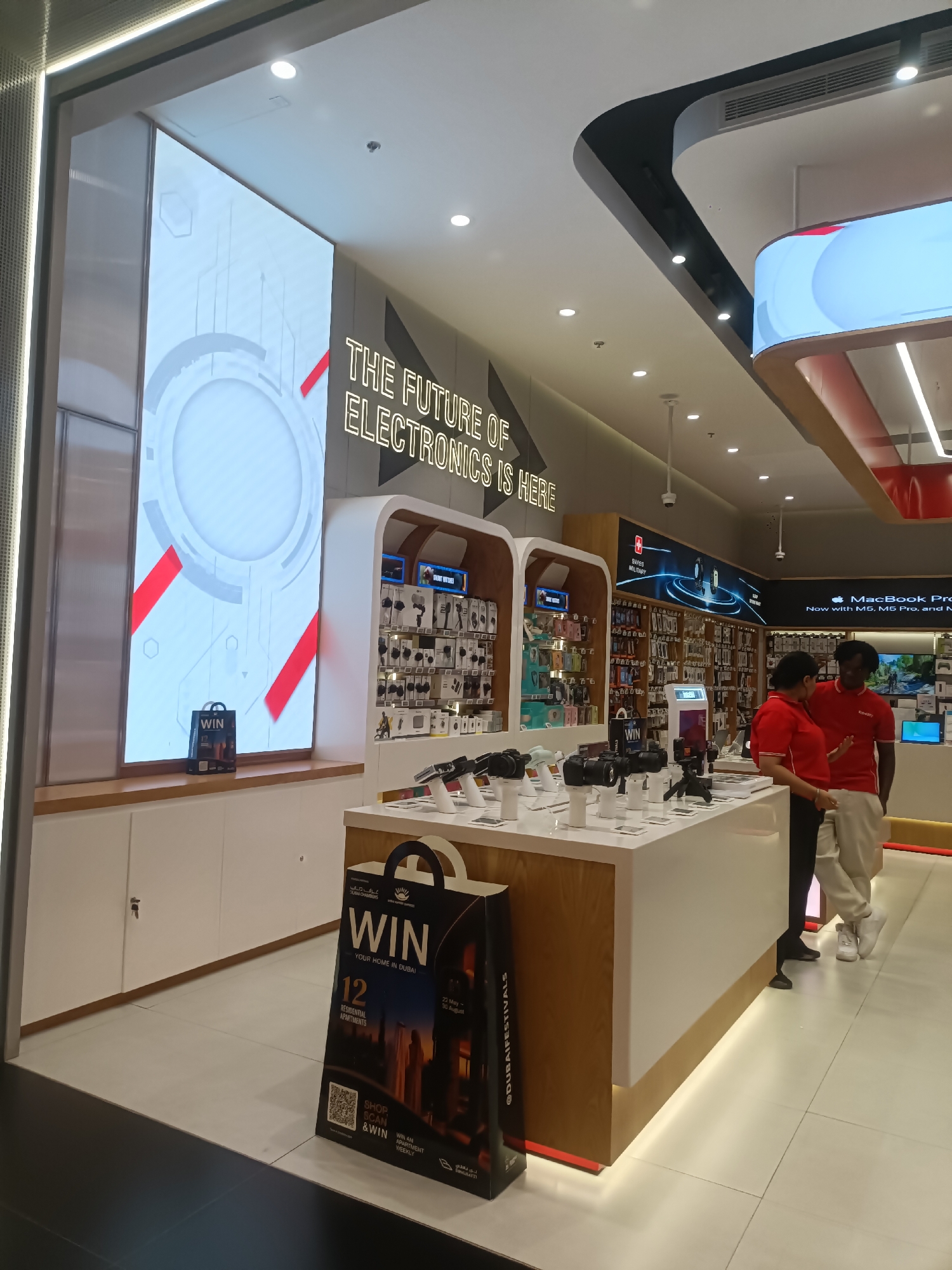 The image depicts the interior of a modern electronics store, likely a retail chain, located in Dubai, United Arab Emirates. The store features a bright, contemporary design with white countertops and shelves showcasing various electronic gadgets. A large LED screen on the left wall displays futuristic graphics and text that reads "THE FUTURE OF ELECTRONICS IS HERE." In the center of the store, two employees, a man and a woman, both wearing red company uniforms, are engaged in a conversation. They stand behind a display counter with cameras and other accessories. A promotional bag with the word "WIN" is prominently placed in the foreground, suggesting a contest or giveaway. The store environment appears clean and well-organized, with ample lighting contributing to its upscale ambiance. The overall impression is that of a high-tech retail space in a bustling urban environment.