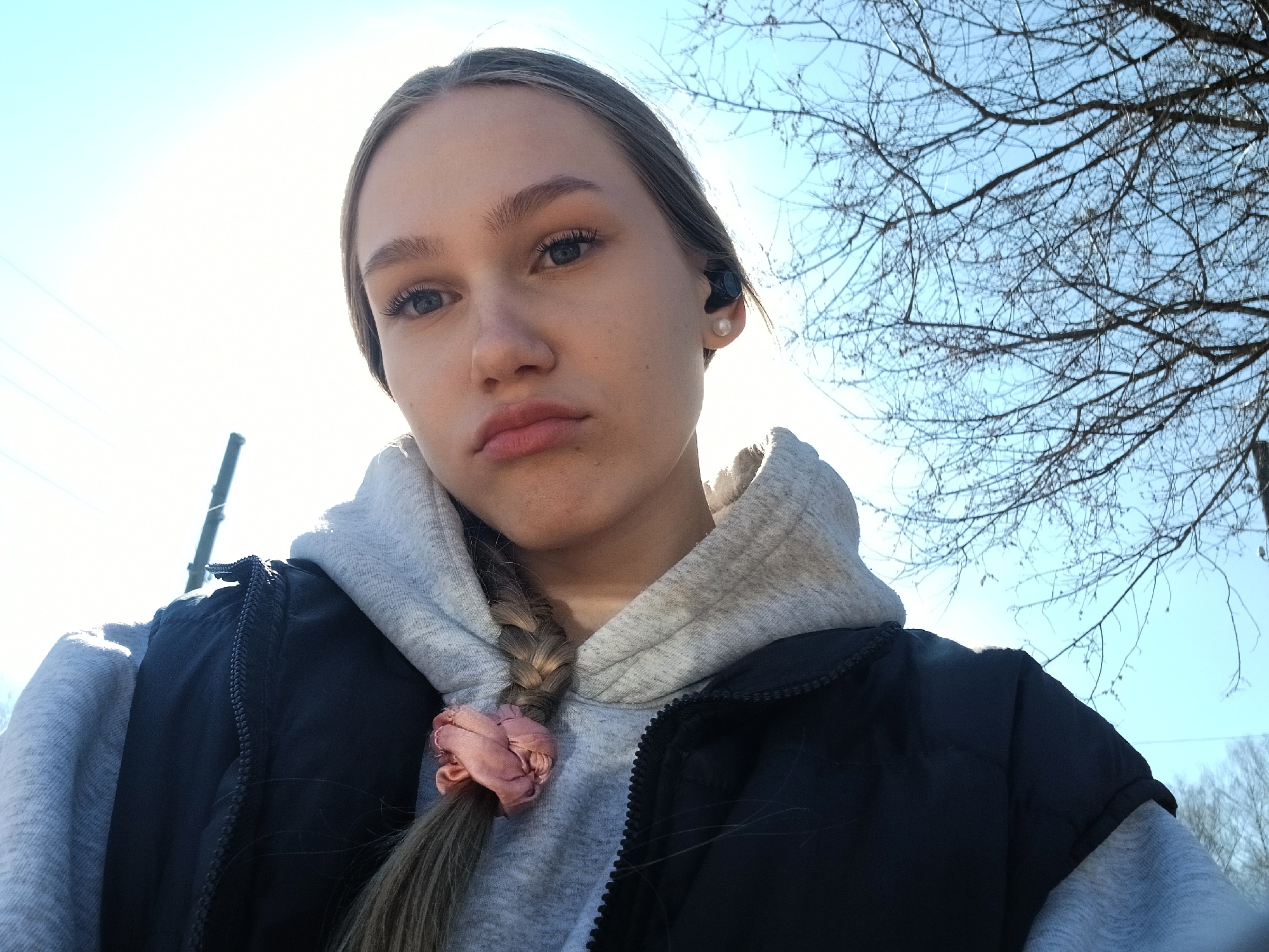 A young woman is captured in a close-up, low-angle selfie, set against a bright, clear sky. She has light brown hair, styled in a braid that rests over her left shoulder, fastened with a soft pink scrunchie. Her gaze is directed at the camera, and her lips form a subtle pout. She is dressed in a light grey hooded sweatshirt, layered under a black padded vest. A black earbud is visible in her right ear, accompanied by a small, white pearl-like earring.
The background features a bright, sunlit sky, transitioning from brilliant white on the left to a light blue on the right. The upper right portion of the frame is dominated by the intricate, bare branches of a deciduous tree, indicating late autumn or winter. A faint, dark vertical line, possibly a utility pole or wire, can be seen in the distant bright area on the far left. The scene suggests a sunny, potentially cool day outdoors in Bolshaya Sosnova, Russia. No text is visible in the image.