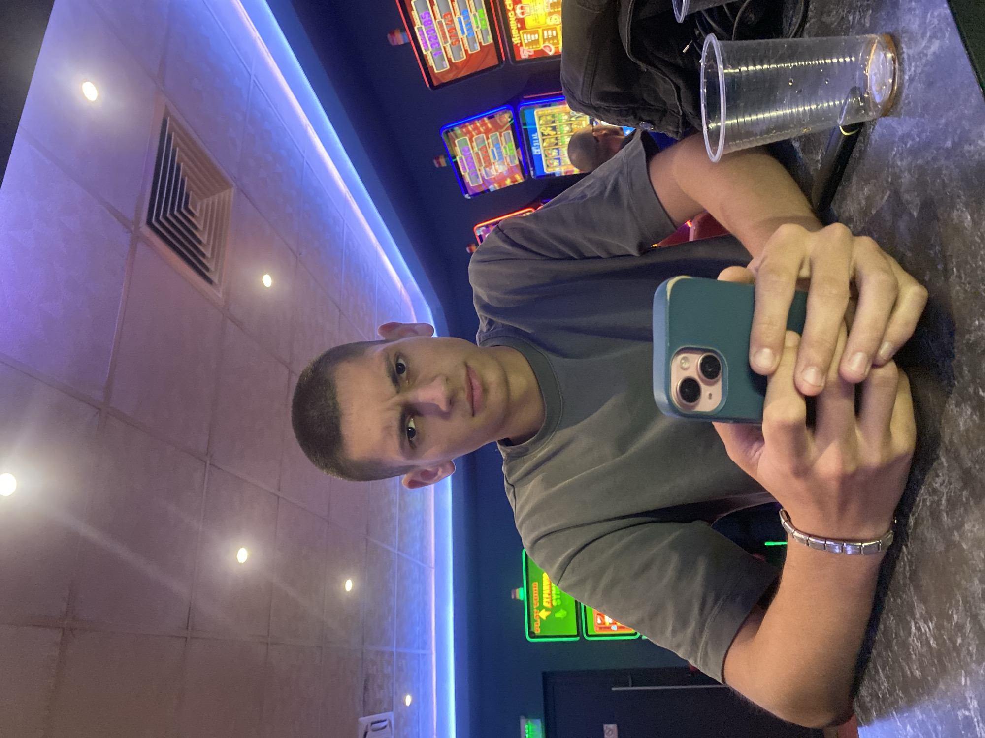 A young man with a buzz cut and a furrowed brow looks directly at the camera while holding a smartphone. He is wearing a dark grey t-shirt and a silver bracelet on his left wrist. He is seated at a dark, speckled counter, and in front of him is a clear plastic cup containing a small amount of amber liquid. The background features several brightly lit slot machines, indicating a casino or similar establishment. A blue neon light runs along the ceiling, which is composed of square panels and has a ventilation grate. The presence of slot machines suggests the location is within the city of Beloslav, Bulgaria, a city known for its entertainment venues.
