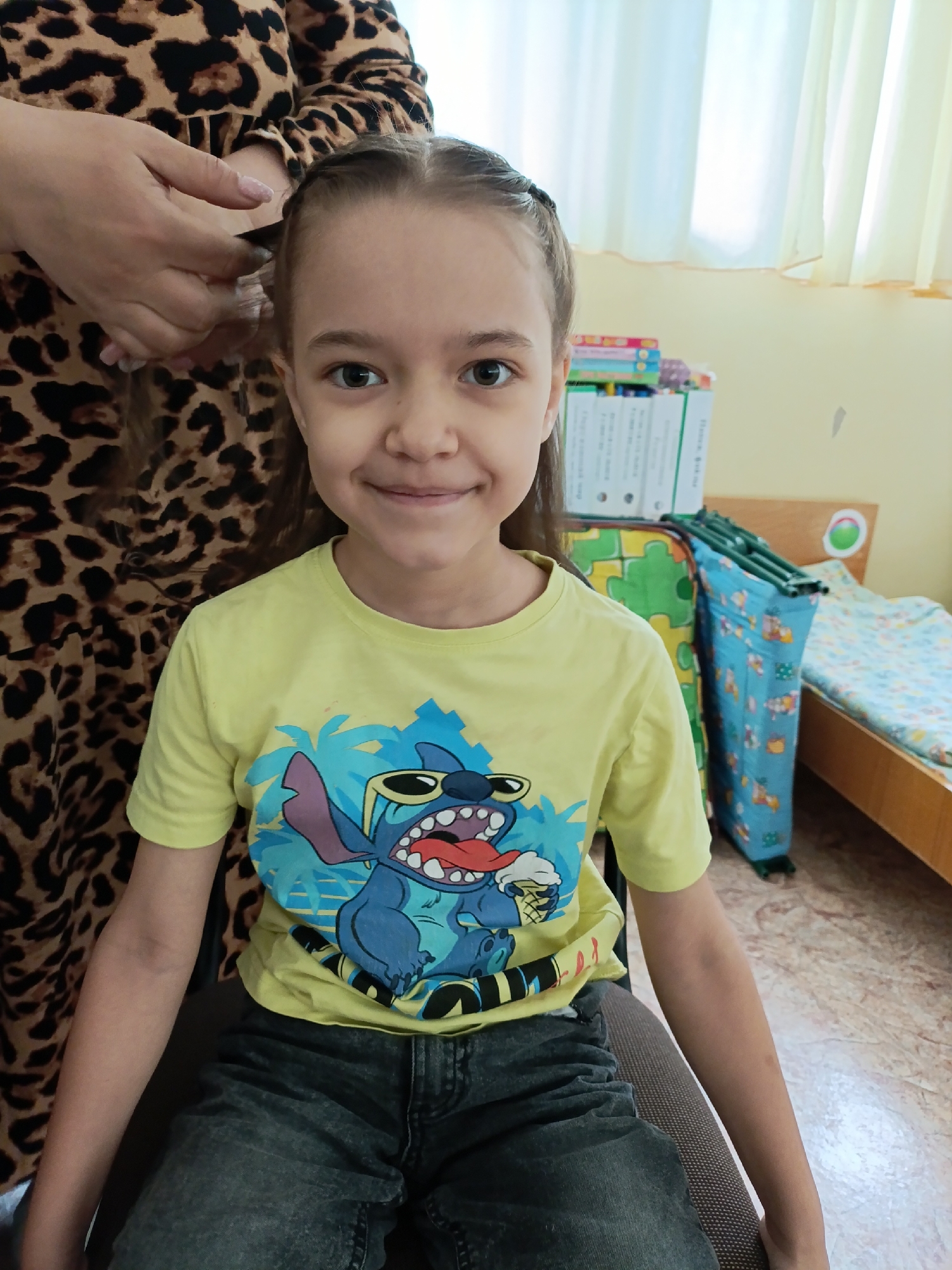 A young girl is sitting on a chair, facing the camera, with a partial view of an adult to her left side who is braiding her hair. The girl has light brown hair, braided partially at the top, brown eyes, and a slight smile. She is wearing a yellow t-shirt with a cartoon graphic of "Stitch" and dark-colored jeans. The adult is wearing a brown and black animal print dress.
Behind the girl, there's a bed, a folded cot with a blue patterned cover, and a stack of books and binders. The room has light yellow walls and a window with light-colored curtains. The floor is tiled in a light marble-like pattern.
Based on the available information, this photo was taken in a residential environment, likely in the city of Chaikovsky, Russia.