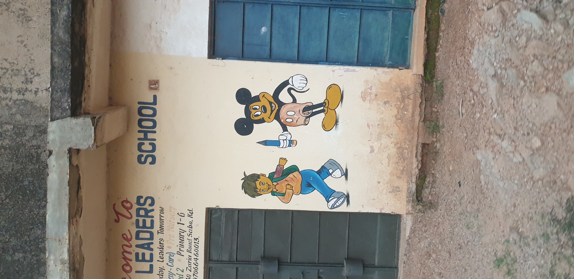 The image shows the exterior wall of a building with faded paint and peeling plaster. A building sign reads "come To LEADERS SCHOOL". Below the sign, the text "Today, Leaders Tomorrow (Day-Care) Pre Nursery" and "Std 2 - Primary 1-6 Zaria Road Sabon, Kad. 07066460013." is visible. To the left of the sign, a faded green metal door is present. To the right of the sign, a dark blue metal door is visible. On the wall, a mural depicts two cartoon characters: a boy carrying a backpack and a pencil, and Mickey Mouse holding a pencil. The ground in front of the building is covered in gravel and dirt with some patches of grass. The location is identified as Sabon Tasha, Nigeria.