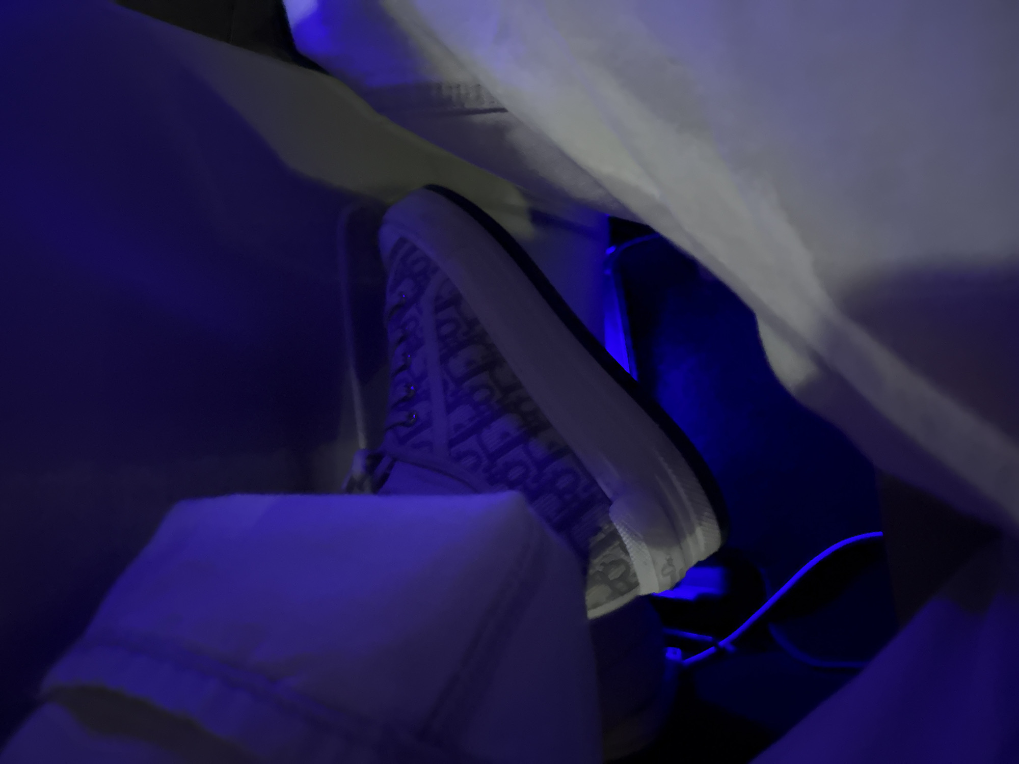 A single foot wearing a Dior sneaker with an oblique pattern is partially visible. The foot is positioned within a space illuminated by strong blue light. White clothing partially covers the scene. A white cable is visible near the shoe. The image lacks context to determine its location in Plovdiv, Bulgaria.