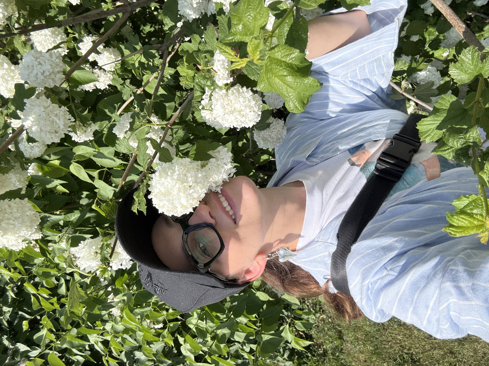 A person with brown hair, wearing glasses, a black baseball cap, a white t-shirt under a blue striped shirt, and a black strap across their body, is lying amongst a bush with white flowers and green leaves. The location is Kazan, Russia. The person is smiling.