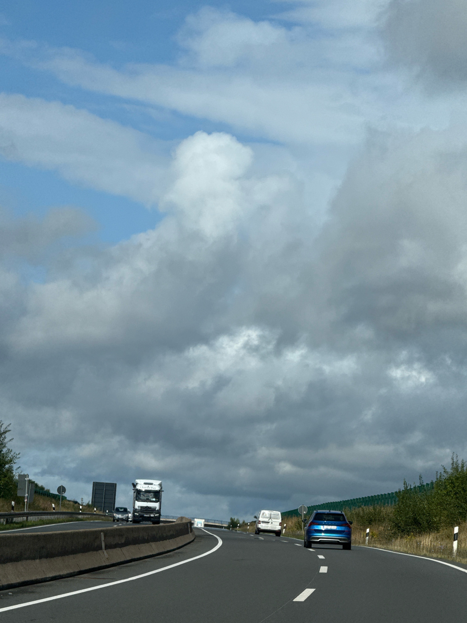 The image shows a view from a moving vehicle on a highway, likely in or near Bad Sachsa, Germany, given the context provided. The sky is filled with a mix of bright blue patches and large, dark gray clouds, suggesting a partly cloudy or overcast day.
In the foreground, the highway has clear white lane markings. A concrete barrier runs along the left side of the road.
Several vehicles are visible on the highway:
- A white semi-truck with "Charity Way" written on its side is in the middle lane.
- A white van is in the right lane, ahead of a blue station wagon.
- The blue station wagon is in the right lane, slightly ahead of the van.
- A small, dark-colored car is visible behind the semi-truck in the left lane.
The sides of the highway are lined with green vegetation, including trees and bushes. On the left side, there are also some road signs and infrastructure, including a dark rectangular structure. A green sound barrier runs along the right side of the highway. The overall impression is a typical highway scene with active traffic under a dramatic sky.