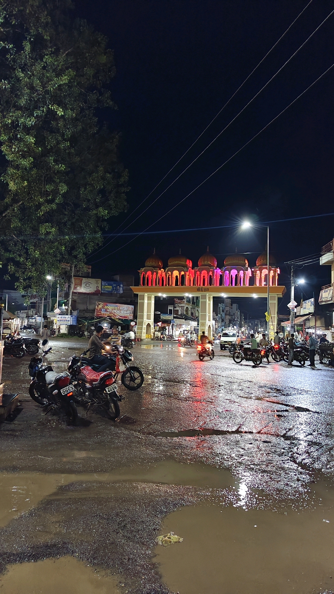 At night, in Thikri, India, an ornate archway decorated with glowing pink and orange lights stands prominently over a wet, reflective street. The archway features multiple domes and intricate pillars. Several motorcycles are parked on the road in the foreground, some with riders. Others are in motion, their headlights cutting through the darkness. The street is covered in puddles, reflecting the ambient light and the colorful illumination from the archway. Buildings line the street on either side, displaying advertisements and illuminated signs. A large tree with dark green foliage is visible on the left side of the image. Overhead power lines stretch across the dark sky. The scene depicts a typical nocturnal streetscape in a town, with signs of recent rain evident on the road surface.