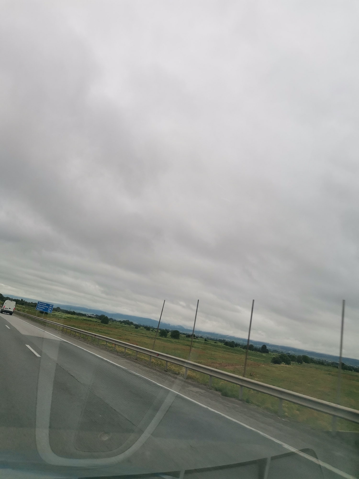 The image shows a view from a vehicle traveling on a highway. The sky is overcast with heavy clouds. On the left side of the road, a white van is visible. A blue road sign with white text is also present on the left, indicating distances to various locations. The right side of the road is bordered by a metal guardrail and a grassy field with some scattered trees. In the distance, hills and a hazy landscape can be seen. The scene suggests a cloudy day in a rural or semi-rural area.