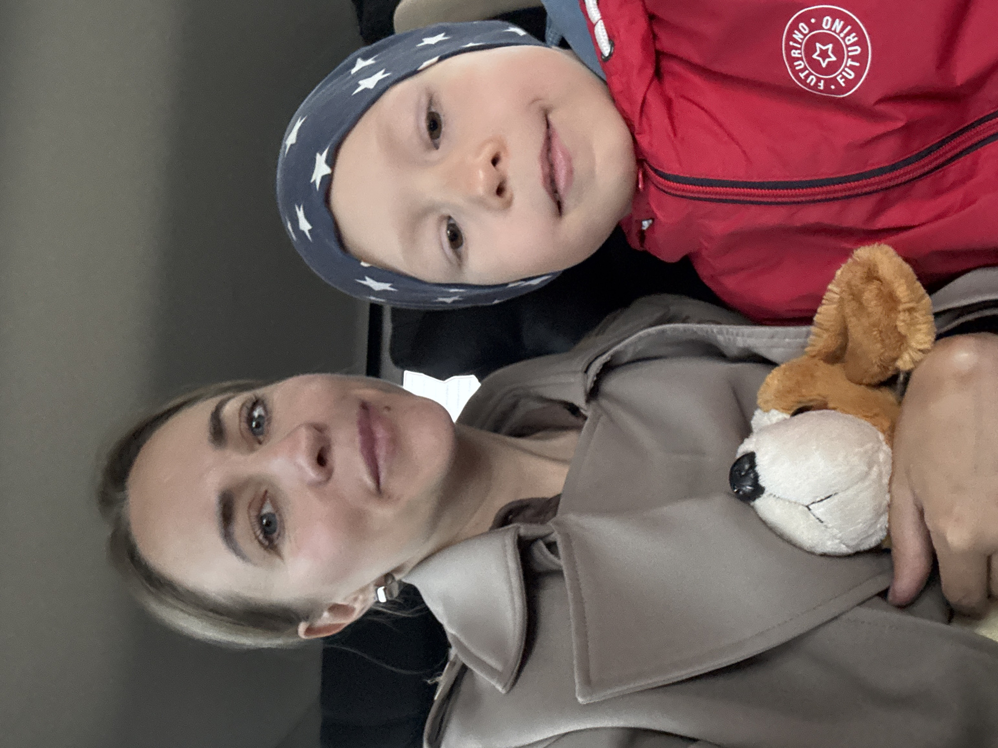 A selfie-style portrait shows a woman holding a young child, likely a baby or toddler, in an indoor setting. The image is oriented vertically, showing the subjects from the chest up against a muted, possibly grey, background.
The woman is on the right side of the frame, looking directly at the camera with light-colored (blue or grey) eyes and a neutral expression. She has light brown hair styled simply. She is wearing a light brown or beige trench coat with a visible collar. Her left hand is visible, holding a plush toy.
The child is positioned above and slightly to the left of the woman, also looking towards the camera with a slight smile. The child has fair skin, brown eyes, and is wearing a dark blue beanie-style hat adorned with white stars. The child is dressed in a vibrant red jacket with a black zippered pocket and a circular white logo on the left chest. The logo clearly reads "FUTURINO" around the perimeter with a star in the center.
The plush toy, held by the woman, appears to be a dog or fox with a white snout, black nose, and brown fur. The indoor setting provides no specific location cues beyond what appears to be a simple wall or car interior. The lighting is soft and diffused, typical of indoor or overcast conditions, and the time of day is not discernible. The overall mood is calm and familial. The location context for this image is Chaikovsky, Russia.