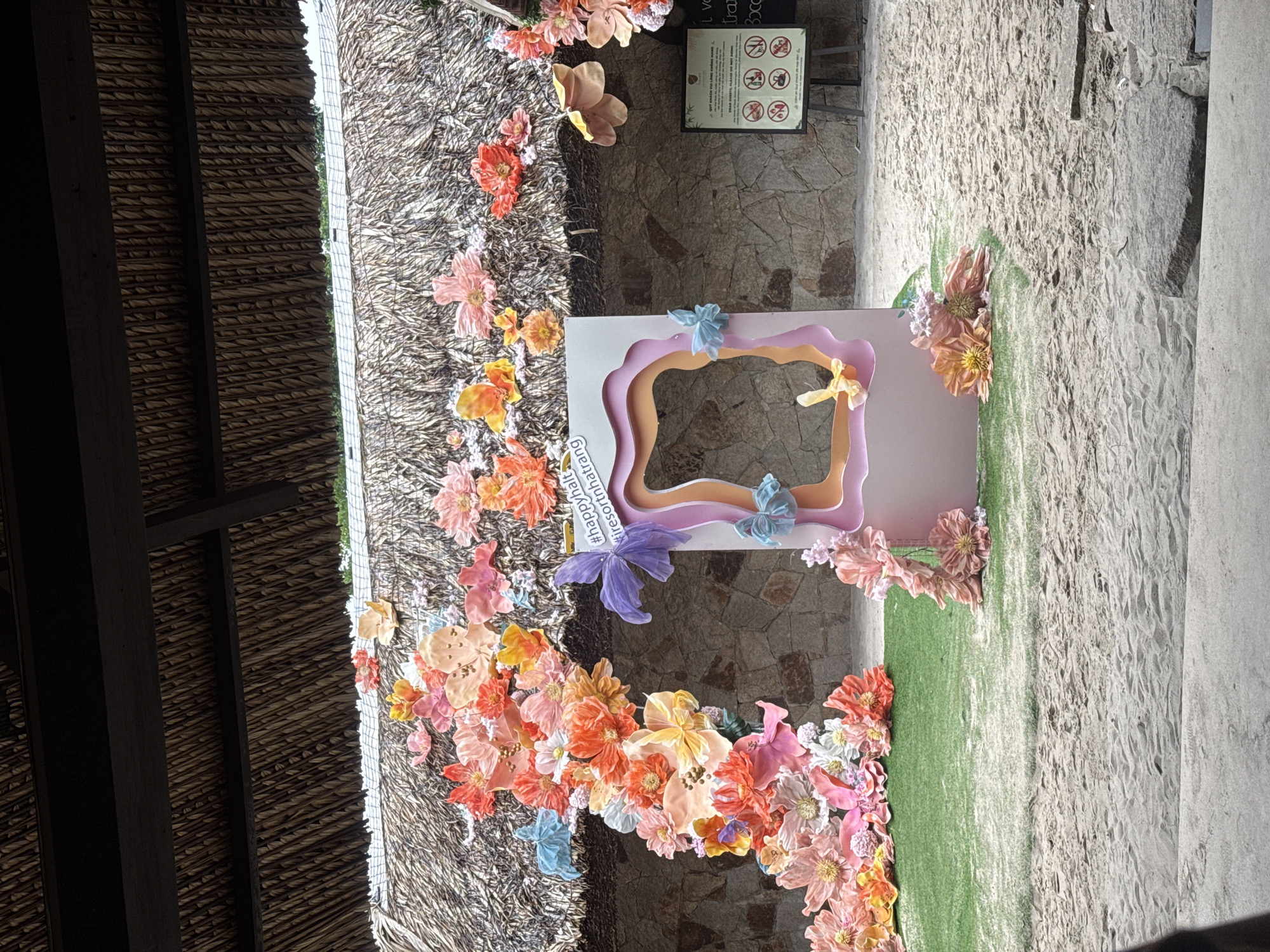 The image depicts an outdoor photo booth decorated with numerous artificial flowers in vibrant shades of pink, orange, and yellow. The photo booth itself is a pastel pink frame with cutouts, adorned with blue and yellow fabric butterflies. Behind the booth, a thatched roof structure provides shade. To the right of the photo booth, a sign with rules in Vietnamese and English is displayed on an easel. The scene is set on a sandy area, with a patch of artificial grass in front of the booth. The background consists of a stone wall. The overall impression is a festive and decorative setup, likely for an event or tourist attraction in Nha Trang, Vietnam.