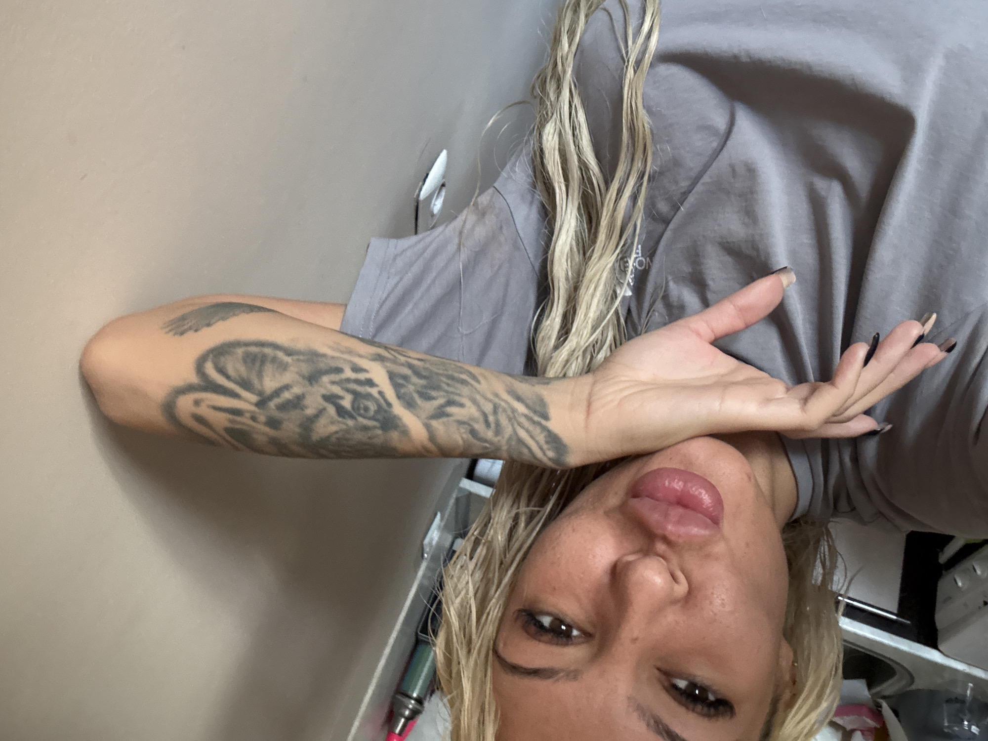 A woman with blonde hair and a tattoo on her arm is positioned in front of a light beige wall. She is wearing a gray T-shirt and looking into the camera with her hand partially obscuring her face. In the background, there's a glimpse of what seems to be a sink or counter area with some objects on it. It is impossible to definitively determine the location in Plovdiv, Bulgaria, based solely on this image.
