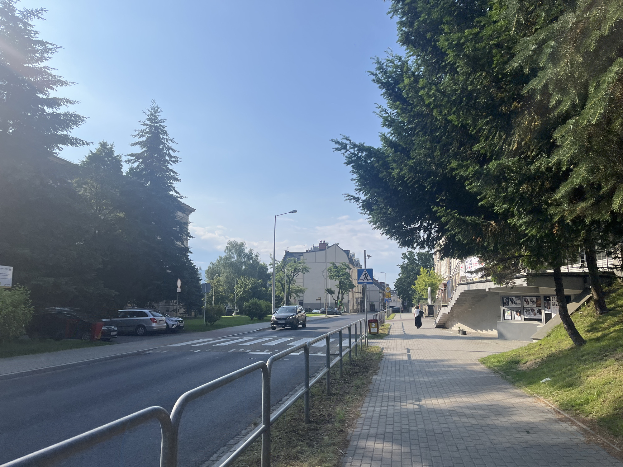 The image depicts a street scene in a city, likely in the daytime given the clear blue sky and sunlight. On the left side of the image, there is a sidewalk with a metal railing separating it from the road. Several cars are parked along the sidewalk, and a few trees line the street. The road itself has a crosswalk marked. Further down the road, buildings are visible, and there are streetlights. On the right side of the image, a sidewalk paved with interlocking bricks runs alongside a grassy slope. A building with an elevated entrance and stairs is present, and posters are displayed on its facade. A person in dark clothing, possibly a woman, is walking away from the camera on the sidewalk. The overall environment appears to be a residential or mixed-use urban area with mature trees and a well-maintained sidewalk.