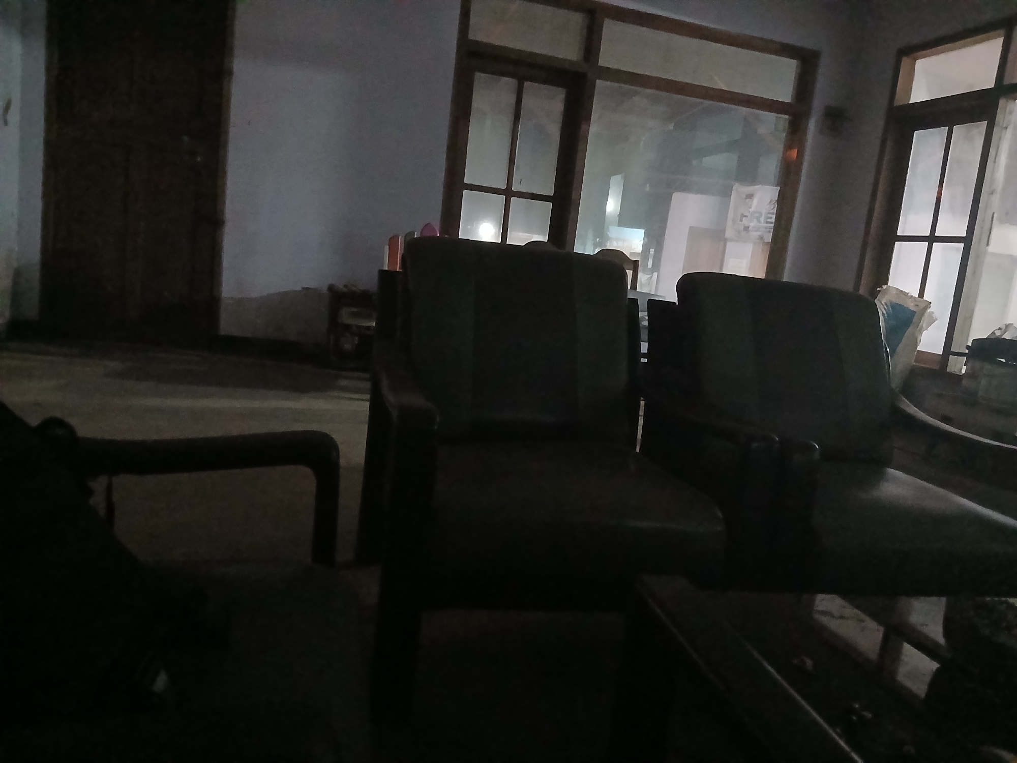 The image depicts an indoor scene, likely a waiting area or lounge, with low lighting. In the foreground, a dark-colored sofa or armchair is partially visible on the left. To the right, two dark-colored armchairs are prominent, facing towards the center. Behind the armchairs, large windows with wooden frames are visible, allowing some natural light and showcasing reflections of the interior. A wooden door is present on the far left of the image. The overall environment appears somewhat dated and possibly under renovation or in disuse, given the dim lighting and general ambiance. No people are visible in the image.