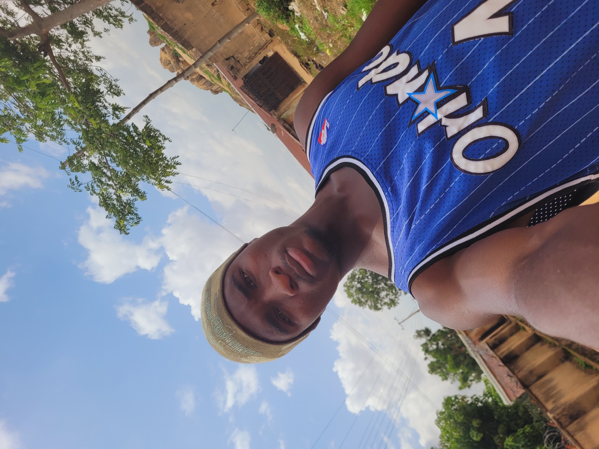 A low-angle, full-body, outdoor selfie shows a Black person wearing a blue Orlando Magic basketball jersey and a light-colored beanie. The person is looking directly at the camera with a neutral expression. Their left arm is extended, holding the camera. The background consists of a bright blue sky with scattered white clouds, a green tree with dense foliage to the left, and some dilapidated buildings and vegetation to the right, suggesting a rural or semi-urban environment in Bukuru, Nigeria. Power lines are visible in the background, crossing the sky. The overall lighting suggests it is daytime.
