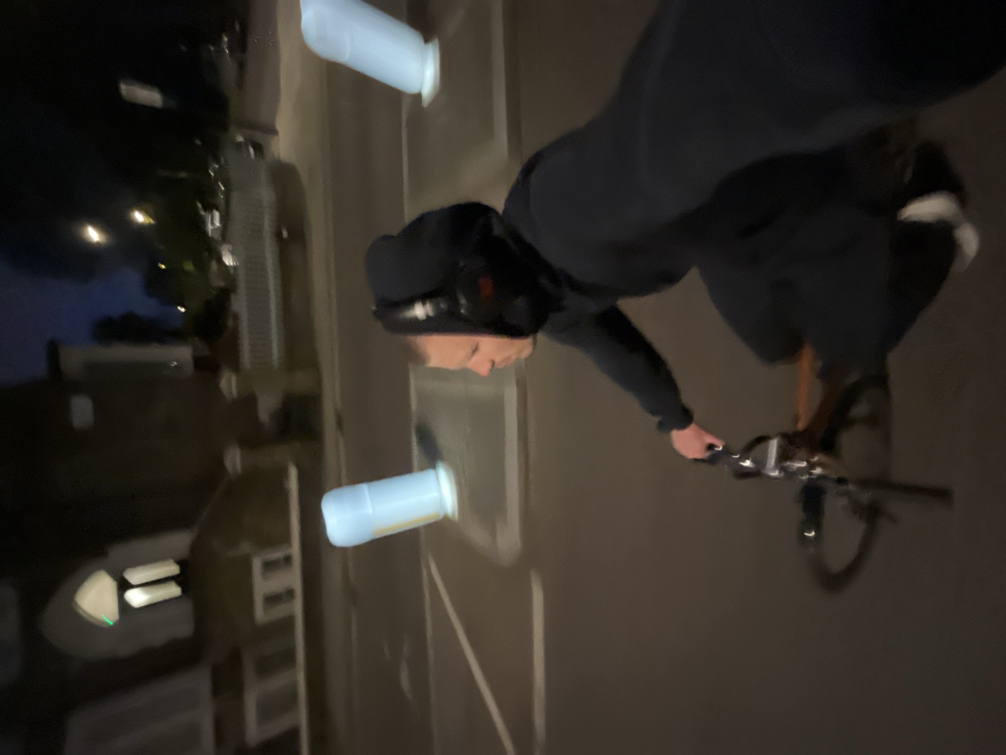 The image shows a person riding a bicycle on a street at night. The person is wearing dark clothing and headphones, and their face is visible. The street is lined with buildings and trees, and there are white bottles on the sidewalk. It is not possible to identify the exact location within the city of London.