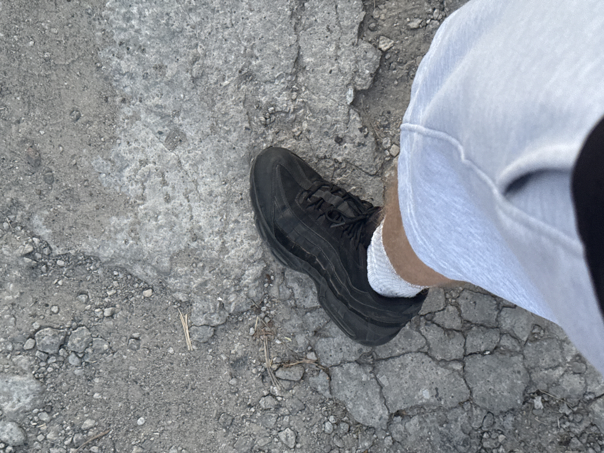 A person's right leg is shown from the thigh down, wearing light blue shorts and a white sock, with a black athletic shoe on their foot. The shoe is positioned on a cracked and uneven asphalt surface. The background consists of the same textured asphalt, with small stones and debris scattered across it. The scene appears to be an outdoor urban environment. No specific landmarks or indicators of the city of Zaporizhzhia, Ukraine are visible.