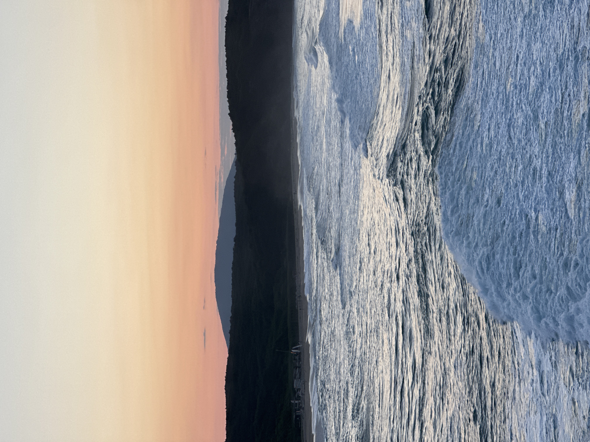 The image depicts a sunset over the Black Sea coast near Sinemorets, Bulgaria. The sky displays a gradient of soft orange and pale yellow hues, transitioning to a lighter tone at the zenith. Silhouetted against the sky are dark, undulating hills covered in dense vegetation. In the mid-ground, a beach is visible with a scattering of figures and what appear to be beach structures or boats. The foreground is dominated by the turbulent sea, with waves cresting and breaking, creating a foamy white texture against the deeper blues and grays of the water. The overall impression is one of a natural, coastal landscape at dusk.