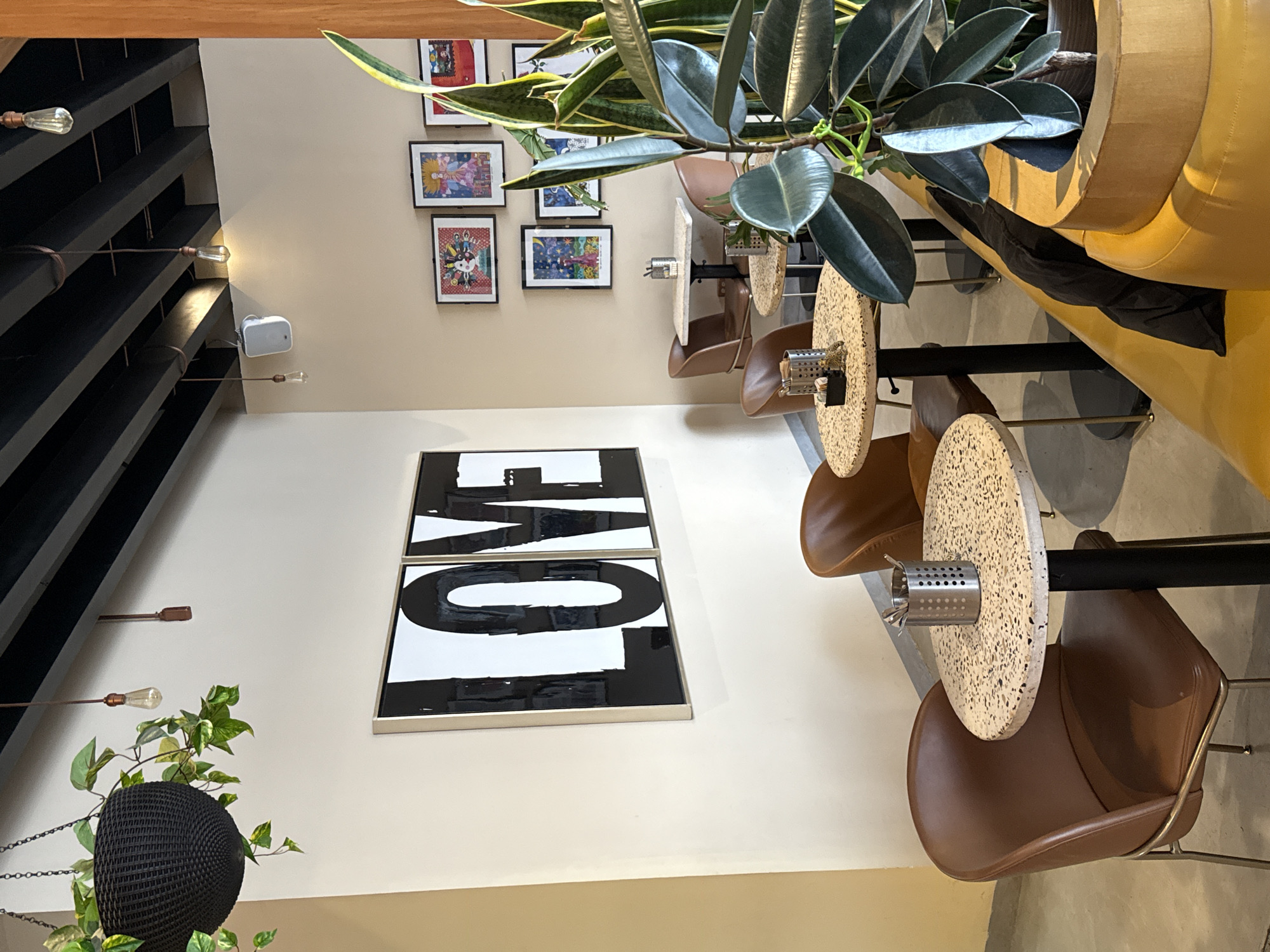 This is an interior shot of a dining area, possibly a restaurant or cafe. The decor includes framed art on the walls, one featuring the word "LOVE" in a bold, modern style. Small round tables with speckled tops are paired with brown leather chairs. Hanging plants and a large potted plant contribute to the ambiance. The ceiling has a distinctive black-and-wood design with exposed light bulbs. While not explicitly stated, the reference to Puebla, Mexico, suggests the image could be from a location within that city.