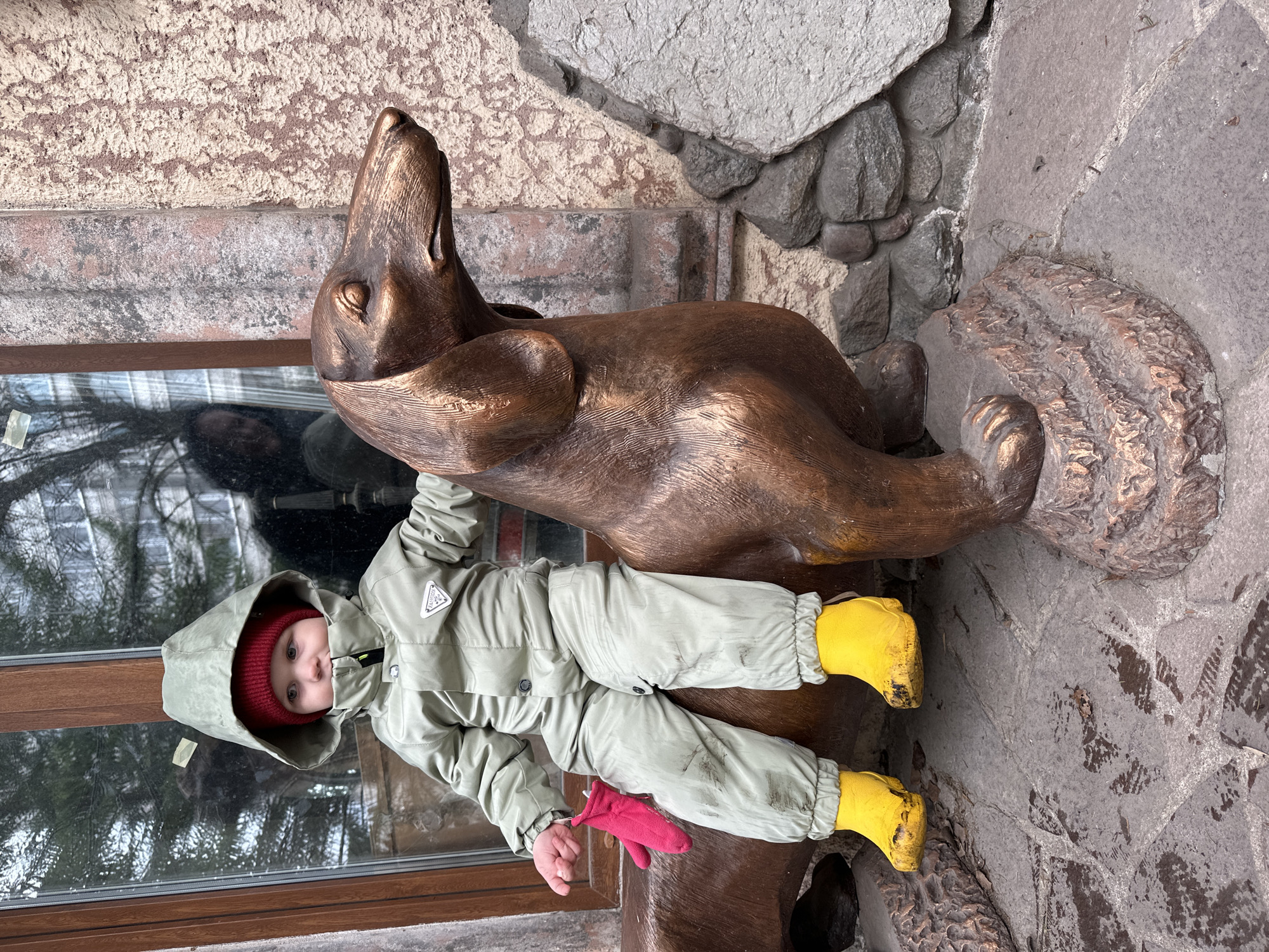 A child wearing a light green snowsuit, a red hat, and yellow boots lies on a bronze sculpture of a seal. The child has a red mitten in their hand. The sculpture is located outside a building with stone facade, likely in the city of Izhevsk, Russia. A window is visible to the left of the child.