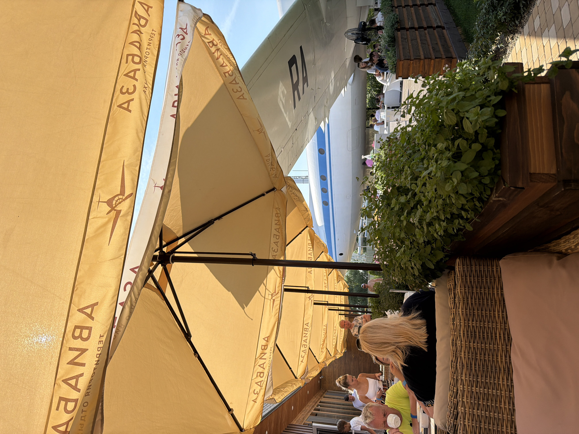 This image depicts the outdoor seating area of a cafe, likely part of the "Abanaba" restaurant complex, located in Kamyshin, Russia. The cafe features a distinctive aviation theme, with an airplane wing prominently visible in the background. Guests are seated at tables beneath several large, light-yellow umbrellas, which provide shade. Some patrons are visible in the foreground and middle ground, engaged in conversation and dining. The overall environment suggests a relaxed, daytime setting with a sunny sky.