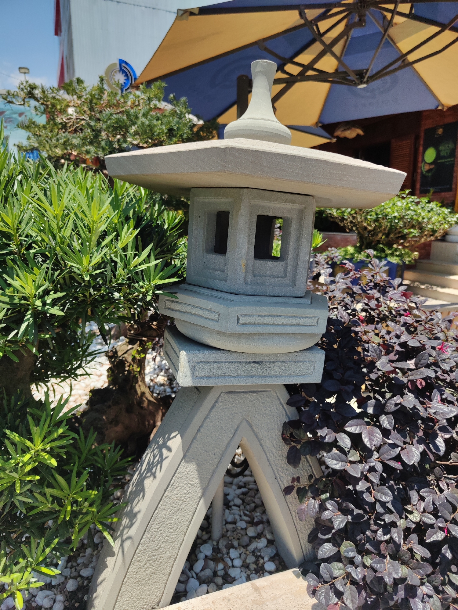 This image shows a stone lantern, traditionally used in Japanese gardens, located in what appears to be a garden setting in Cam Ly - Đà Lạt, Vietnam. The lantern is gray, composed of a base with arched supports, a middle section with a square housing containing window-like openings, and a multi-tiered roof topped with a decorative finial.
The lantern is surrounded by various plants and shrubs, including a green leafy plant to the left and a dark, purplish-leaved plant to the right. Small white pebbles cover the ground around the base of the lantern. In the background, an umbrella with alternating blue and yellow panels is visible, along with glimpses of a building. The scene is bathed in natural sunlight, indicating a daytime setting.