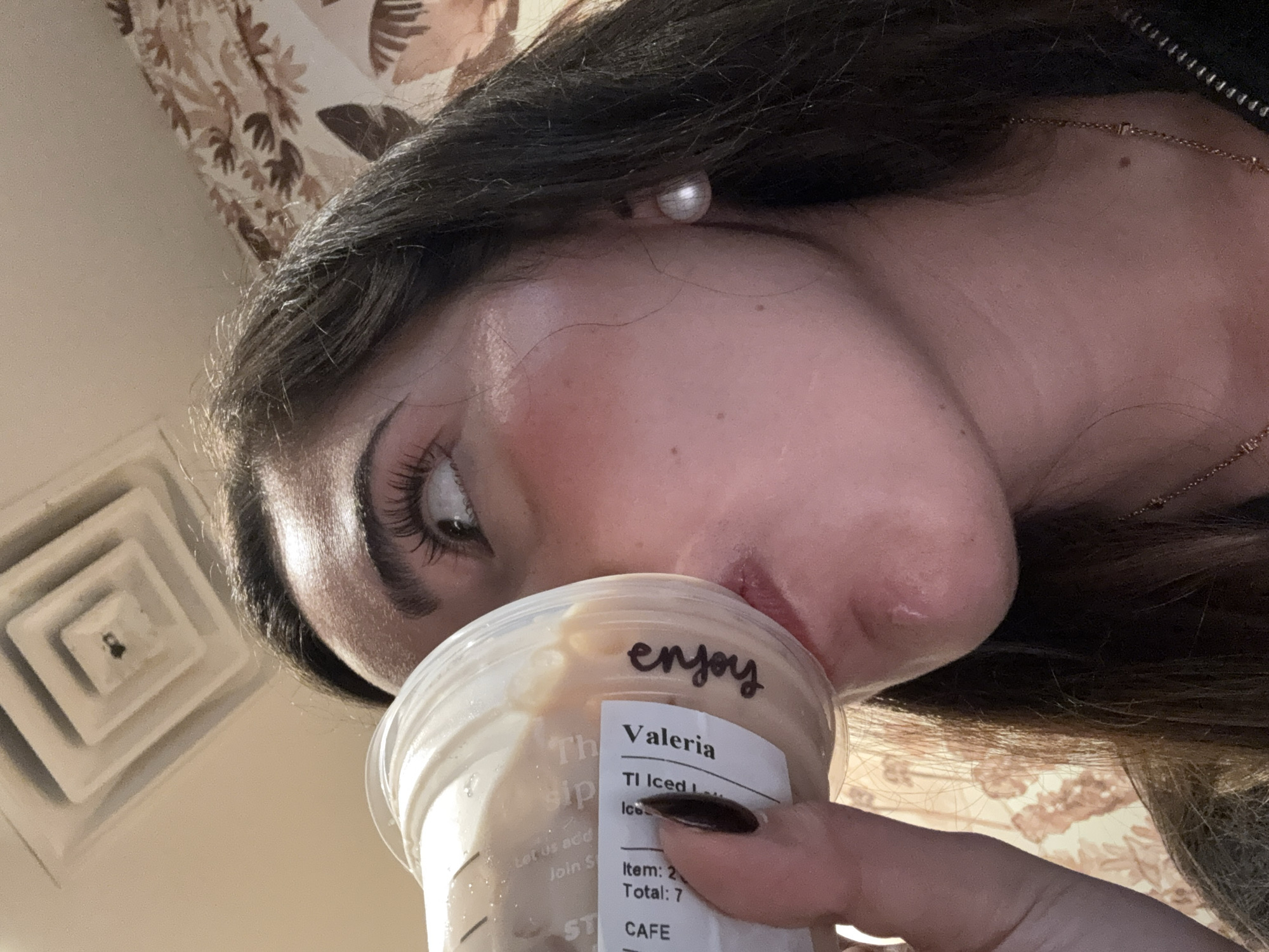 Here is a forensic description of the image:
**People:**
* One person is visible in the image, a woman named Valeria based on the coffee cup label. She has dark hair, defined eyebrows, and visible eyelashes. She is wearing a pearl earring and a necklace.
**Subjects:**
* The primary subject is the woman, partially visible from a low-angle perspective.
* A Starbucks coffee cup with the name "Valeria" and the word "enjoy" written on it.
**Objects:**
* Starbucks coffee cup with a label.
* Pearl earring
* Necklace
* Air conditioning vent on the ceiling
* Fingernails with dark nail polish
**Actions:**
* The woman is holding the coffee cup and about to drink from it.
**Interactions:**
* The woman is interacting with the coffee cup.
**Environment:**
* The environment appears to be indoors, as suggested by the ceiling, air conditioning vent, and patterned curtains/wall covering.
* The location is identified through the context of the prompt as San Antonio, United States of America.