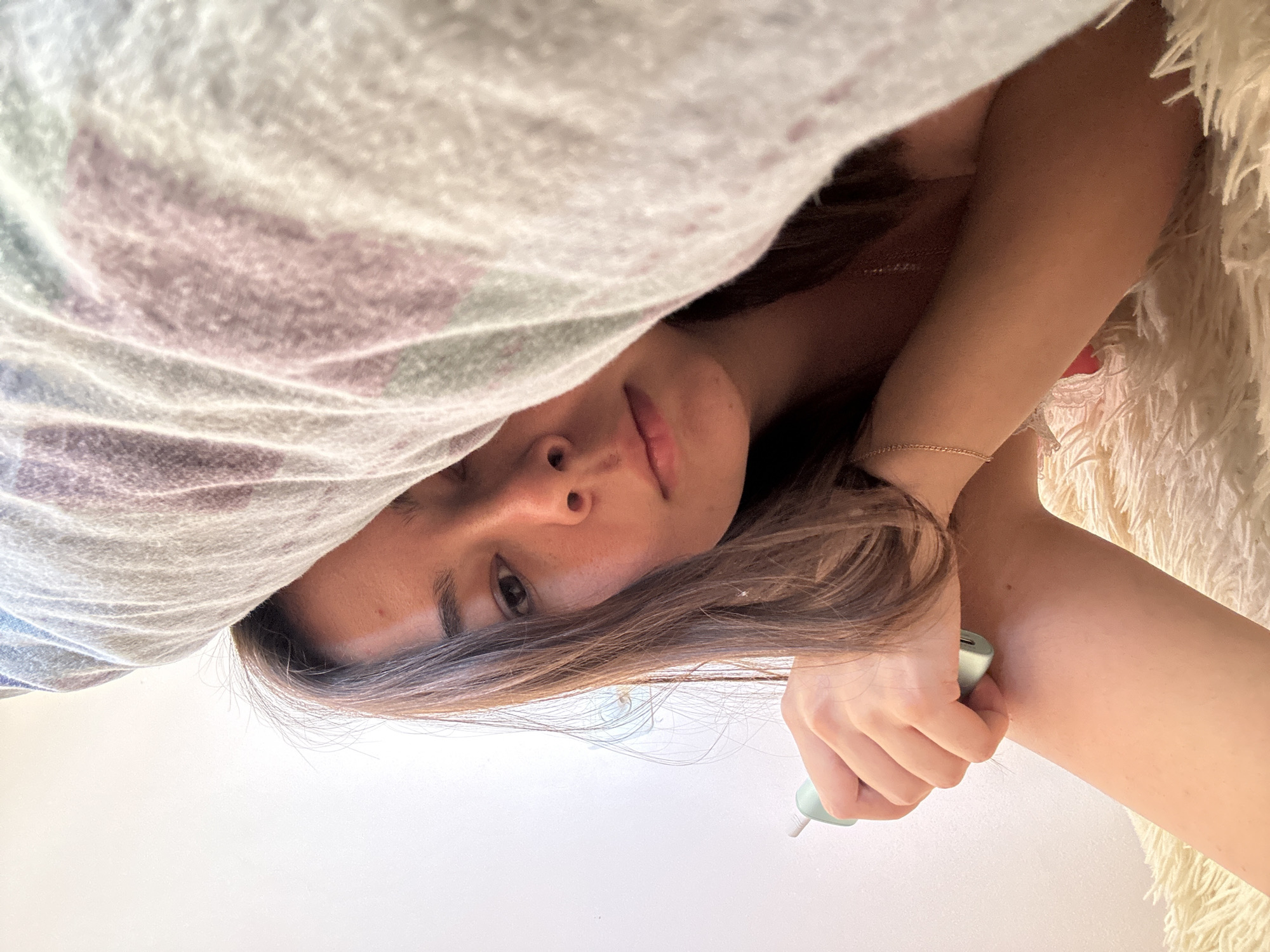 A young woman is lying under a blanket, her face partially obscured. She is holding a light blue device in her right hand. The blanket has a striped pattern in shades of pink, purple, and white. She is lying on a fluffy, white surface. The background is a plain white wall. There is no identifiable information in the image that could place it in Gyumri, Armenia or any other specific city.