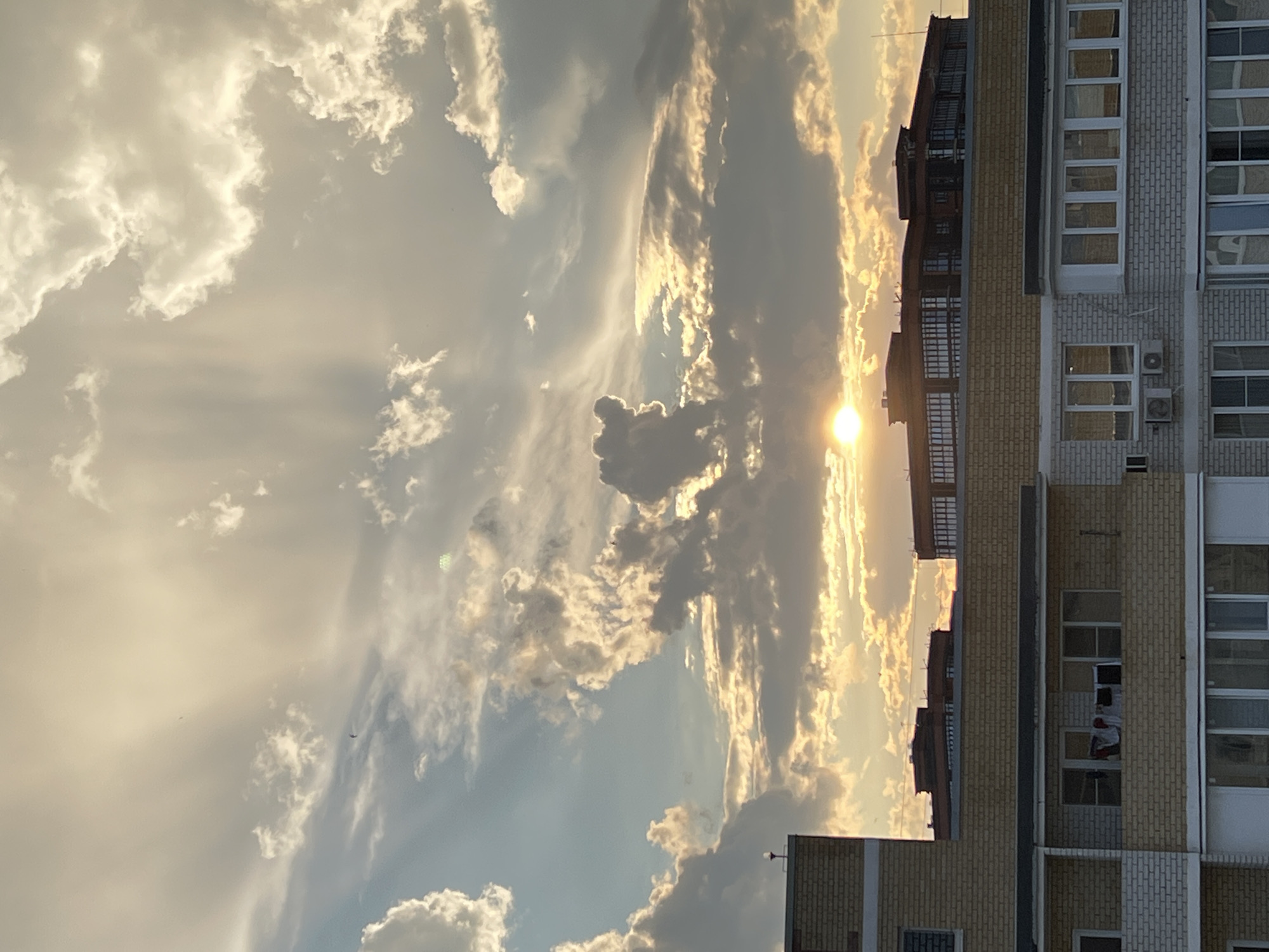 This is a vertical, outdoor, daytime photograph taken in Krasnodar, Russia. The image features a dramatic sky filled with clouds illuminated by the setting sun. The sun, a bright orb of light, is visible near the center of the frame, casting golden rays through the clouds. The sky transitions from bright yellow and orange near the sun to lighter blues and grays on the left side of the image.
To the right of the sky, a multi-story brick building occupies the right half of the frame. The building has numerous windows, some of which reflect the sky. Visible on one of the balconies, laundry is hanging out to dry, including what appears to be red and white clothing. Some air conditioning units are also visible mounted on the exterior of the building.
The overall scene evokes a sense of warmth and calm, typical of a sunset. The presence of the building grounds the image, indicating an urban or suburban setting. The weather appears to be clear with scattered clouds, creating a beautiful visual display as the sun begins to set. There is no visible text in the image.