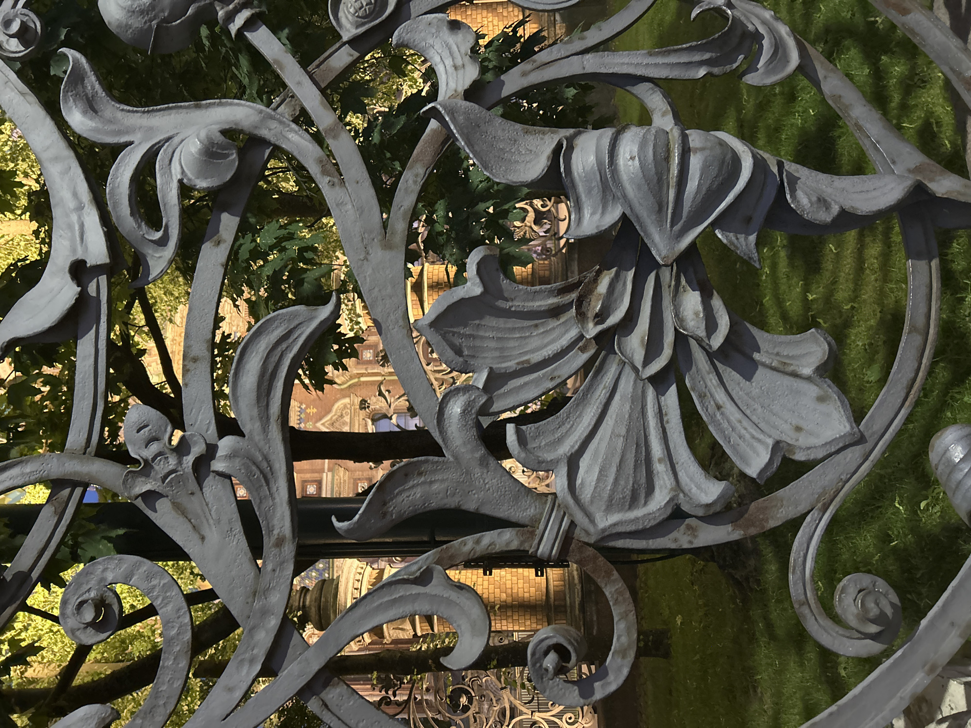 A close-up, eye-level shot captures the intricate detail of a weathered, grey wrought-iron fence in front of a building. The fence features ornate floral motifs and swirling curlicues, with visible signs of rust in some areas. Behind the fence, a lush green lawn extends towards the building. The building itself appears to be constructed of brick and is partially obscured by the shadows of trees. The building's facade is detailed with architectural ornamentation, including what appears to be mosaics or decorative tiles, illuminated by warm, artificial lighting. The lighting suggests it is either dusk or night.