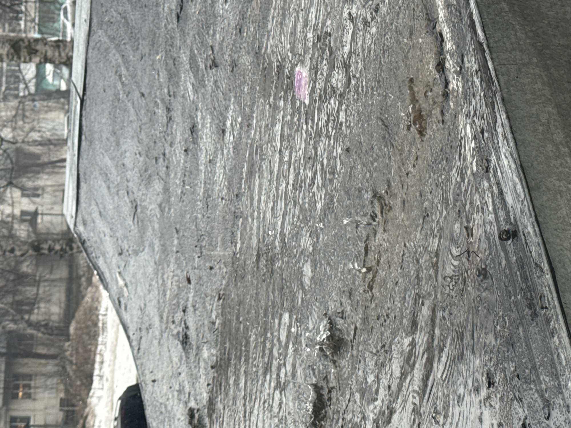 The image shows a close-up of a textured gray surface, possibly a stone or concrete structure. The texture appears fluid-like with vertical streaks. In the background, out of focus, are buildings and bare tree branches. There's a small purple mark on the gray surface. The location is potentially Chaikovsky, Russia.