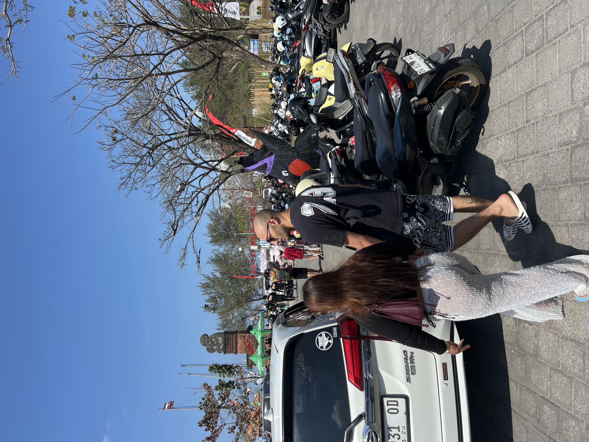 An exterior, eye-level, medium shot shows a couple walking away from the camera on a paved area with a large number of parked motorcycles and a white car on the left. The man, with a shaved head and sunglasses, wears a black t-shirt with white numbering and black shorts with a white pattern. The woman, with long brown hair, wears a black long-sleeved top and translucent white pants. The white car has a license plate reading "31 OD" with a sticker on the rear door. The motorcycles are predominantly scooters and are parked in a dense formation. In the background, there are trees, some tents, and a brick structure with a statue on top. A flag is visible on a flagpole to the left. The sky is clear and blue. This scene is consistent with a public area in Denpasar, Indonesia, potentially near a beach or event.
