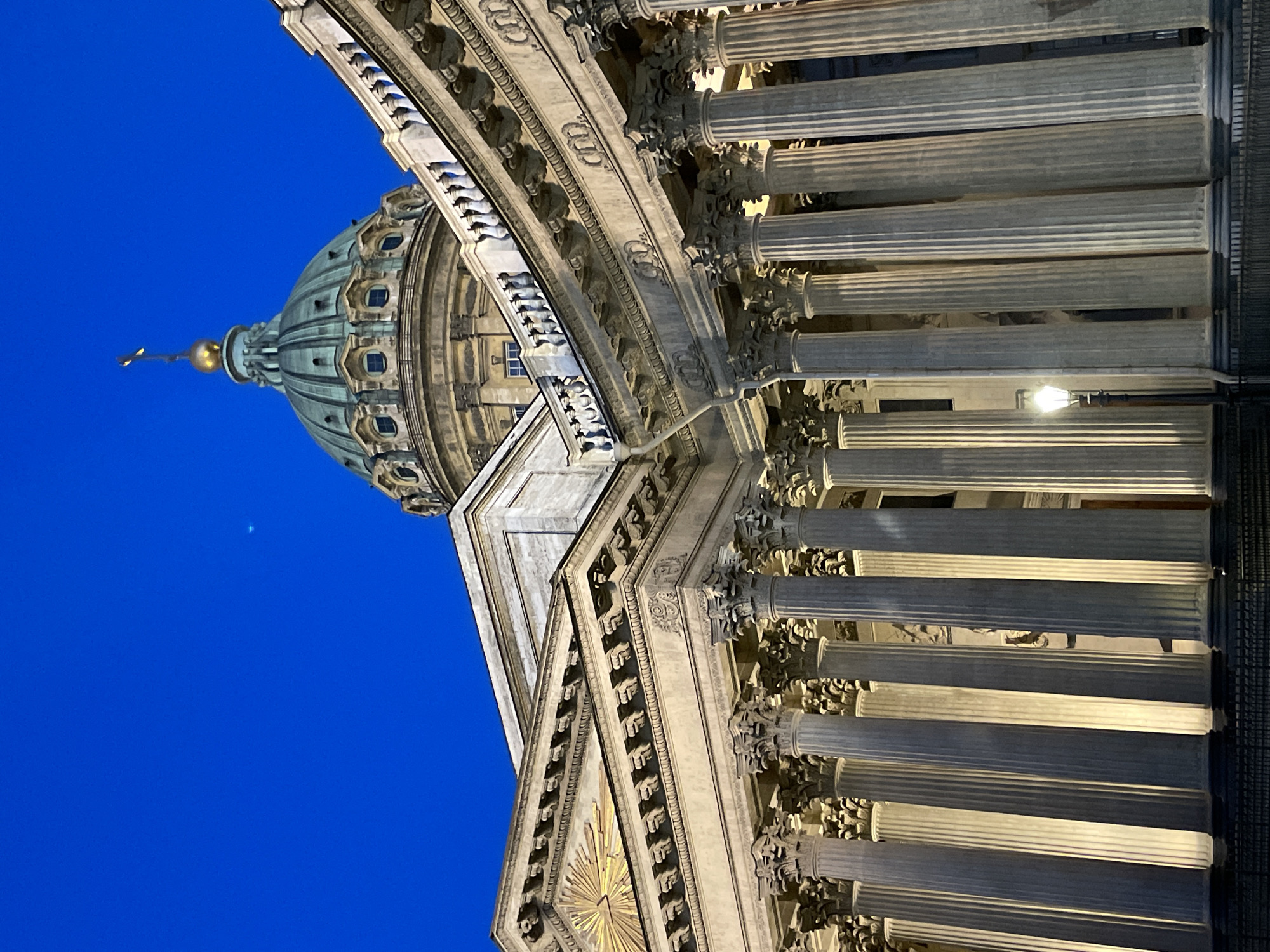 The image depicts the grand Neoclassical architecture of Kazan Cathedral in Saint Petersburg, Russia, viewed from a low angle. The dominant features are the cathedral's large dome and its sweeping semicircular colonnade.
The majestic, greenish-bronze dome, likely oxidized copper, rises prominently against a deep blue sky, suggesting either dusk or very early morning. It is topped with a golden finial and a cross or spire. Below the dome, the main facade of the building is visible, featuring intricate stone carvings, cornices, and a pediment adorned with a radiating sunburst-like emblem.
The colonnade comprises numerous tall, fluted columns with ornate Corinthian capitals, which are artificially lit, casting shadows and highlighting their architectural details. A modern streetlamp is visible within the colonnade, further indicating artificial illumination. The sky is clear, with no clouds. No people or activities are visible, focusing solely on the architectural grandeur of the landmark. The monumental scale and intricate detailing of the structure are emphasized by the upward-looking perspective. No text is visible in the image.