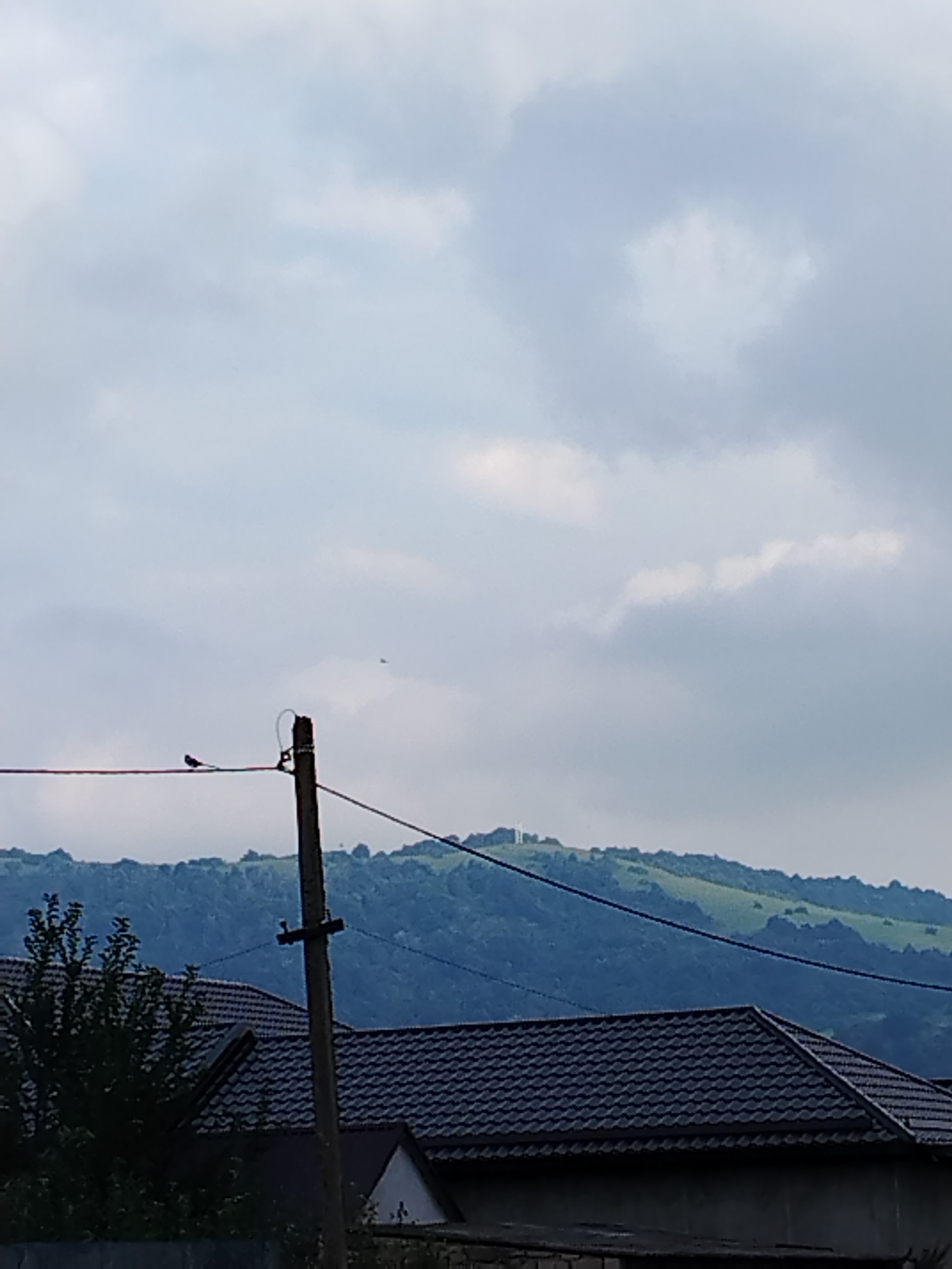 The image shows a natural landscape with a cloudy sky. In the foreground, there are roofs of houses and a utility pole with wires. A small bird is perched on one of the wires. In the background, there is a green, hilly landscape, possibly a mountain range, with a white structure visible on the top of one of the hills. There is also a distant object in the sky, possibly an aircraft. The scene is likely located in a rural or suburban area, possibly in the vicinity of Атажукино, Russia, given the context.
