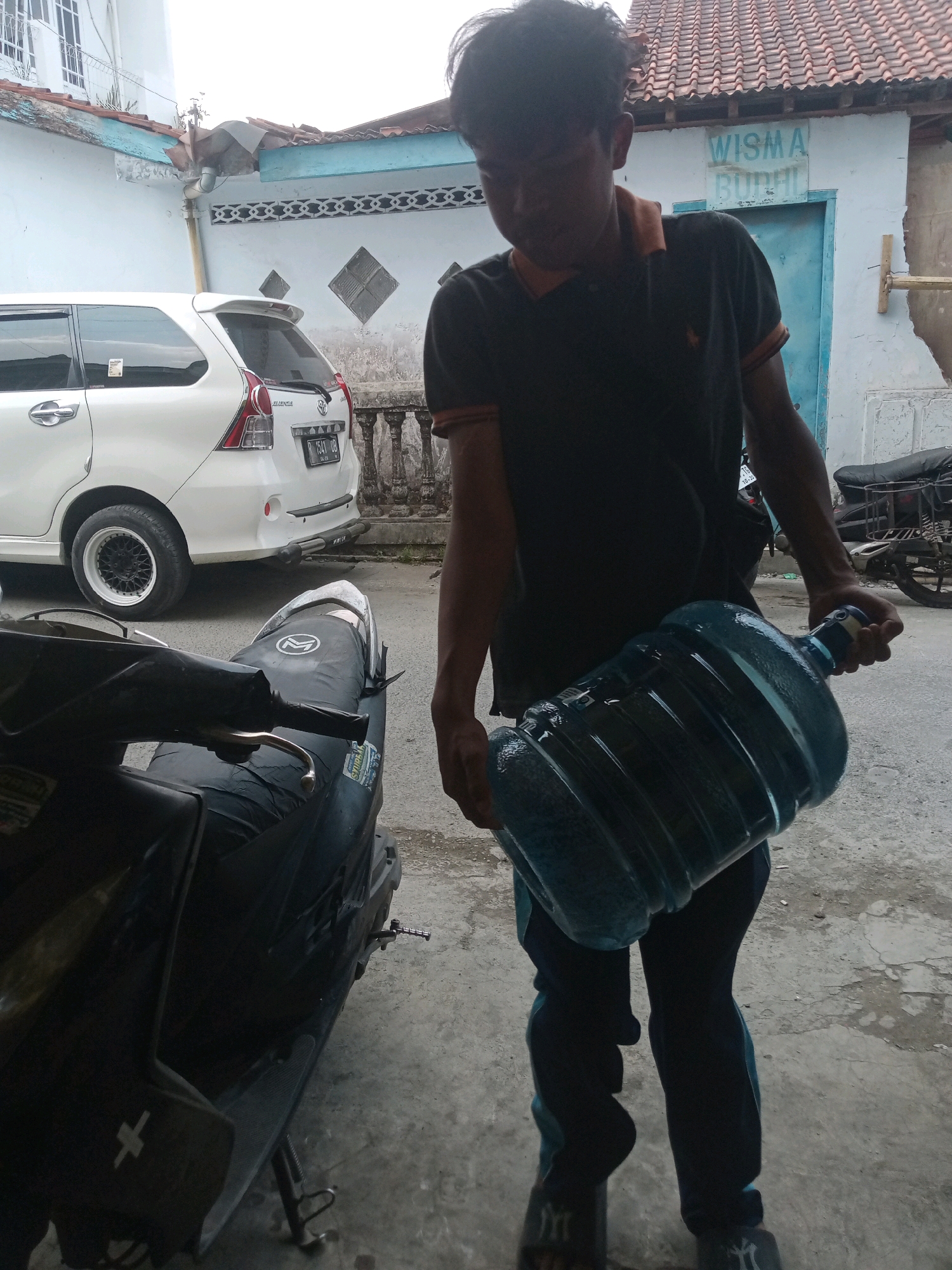 In the city of Grendeng, Indonesia, a young man is depicted carrying a large, translucent blue water jug. He is wearing a dark polo shirt with an orange collar and dark pants with blue accents. The man is standing on a concrete surface, possibly a driveway or street, in front of a white building with a red-tiled roof. A white van is parked to his left, and a black motorcycle is visible in the foreground to his left. The building in the background has a sign that reads "WISMA BURUH". The scene appears to be outdoors, with natural lighting.