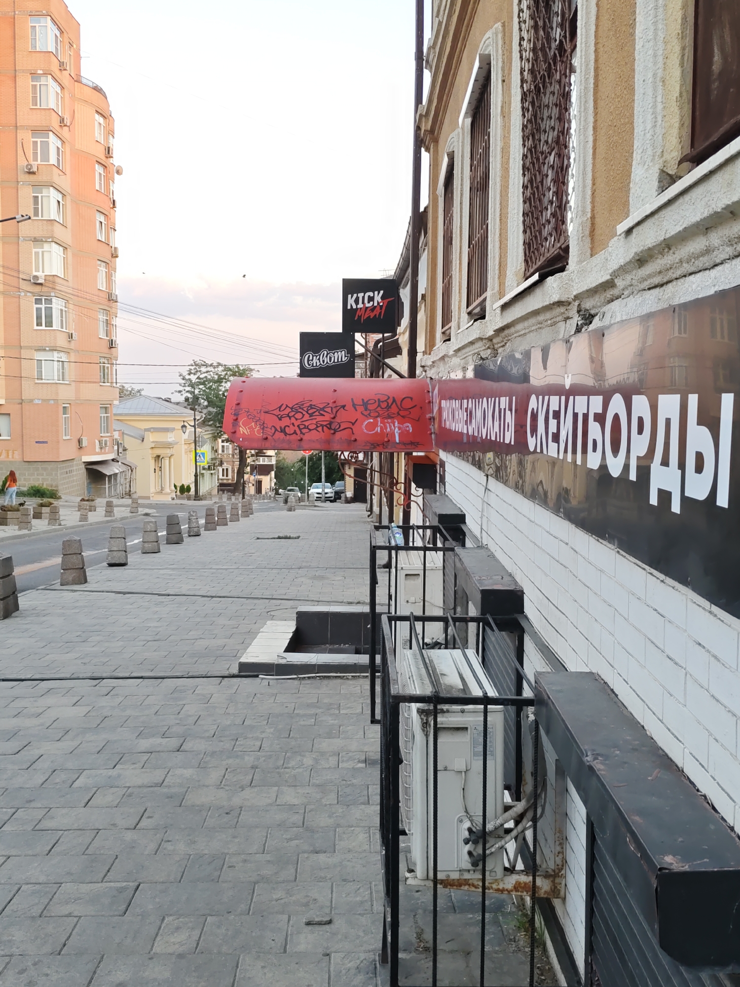 This image depicts a street scene in Rostov-on-Don, Russia. On the left, a tall, light-colored apartment building with many windows and air conditioning units is visible. The street in front of it is paved with rectangular grey stones and lined with concrete bollards. On the right, a building with a textured light-colored facade features barred windows and a large black banner with white Cyrillic text. Below this banner, a red awning is draped, also with graffiti. Several outdoor air conditioning units are mounted on the building's exterior, enclosed by black metal bars. The sky is overcast and a few birds are visible. The scene appears to be a commercial street with shops.
