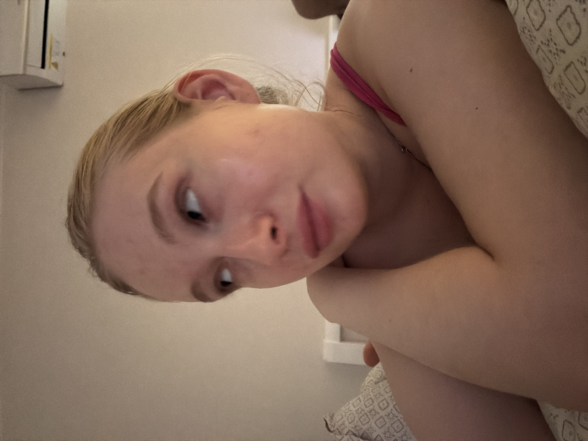 A fair-skinned person with blonde hair is lying in bed, angled towards the camera. They are wearing a pink tank top. The background shows a light-colored wall and a white air conditioning unit. A patterned pillow is visible beside them. The location is Rossiyskiy, Russia.