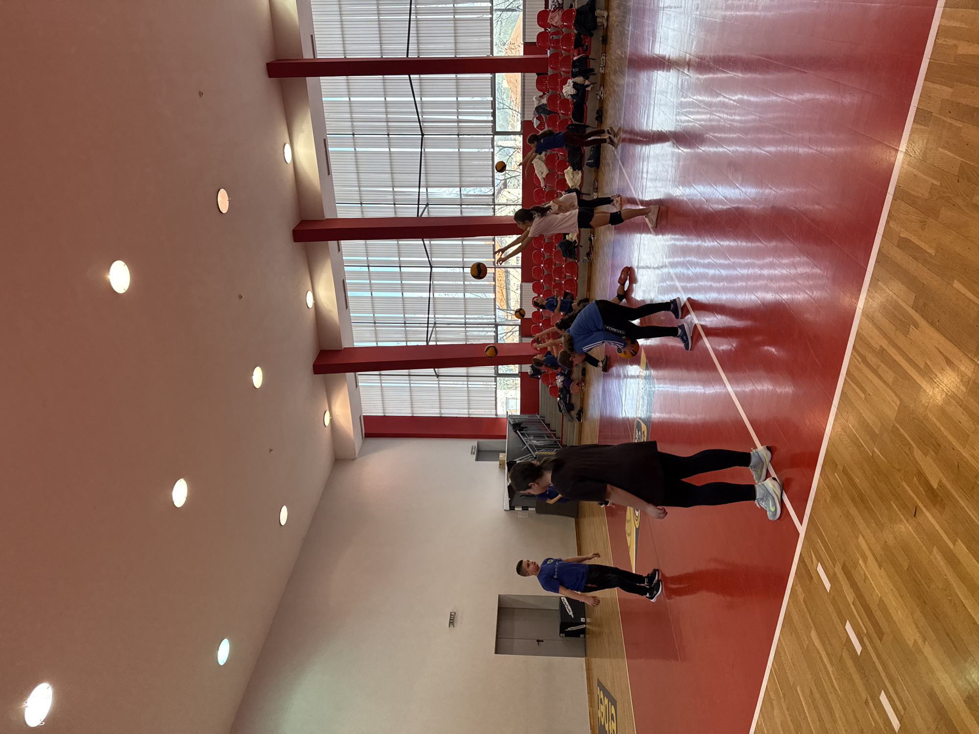 The image shows the interior of a gymnasium in Etropole, Bulgaria. A group of people are lined up on a red basketball court, some holding basketballs and reaching out with their hands as if practicing a drill. Red chairs are visible near the back of the gymnasium. The walls are a light color with embedded lights, and the ceiling has a skylight structure. A man in a blue shirt stands near a doorway on the side of the court. The flooring appears to transition from red to wood on one side.