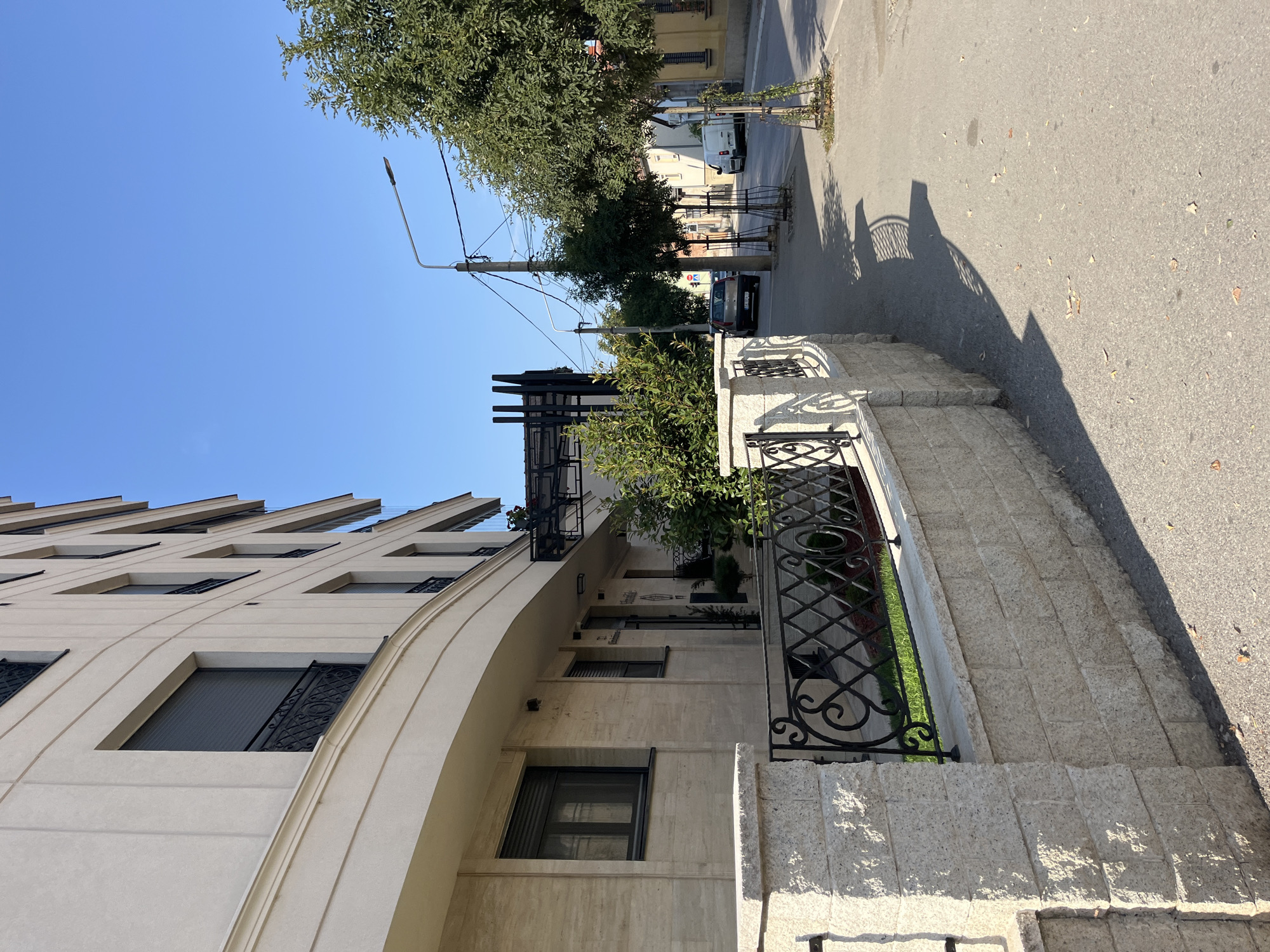 The image depicts a street scene in the city of Cacak, Serbia, on a clear, sunny day. In the foreground, a light-colored stone wall with a decorative black metal fence borders a sidewalk. Beyond the wall, a modern, multi-story building with light-colored facade and dark-shuttered windows is visible. The building has a curved section at the front.
To the right of the building, a sidewalk runs along a paved street. Several trees are planted along the sidewalk, some with protective cages around their bases. Utility poles with electrical wires and streetlights are present along the street. A dark-colored car and a white van are parked on the street. The shadow of the fence and the building are cast across the sidewalk and street, indicating the direction of the sun. The environment appears to be a residential or commercial area with a clear blue sky overhead. No people are visible in the image.