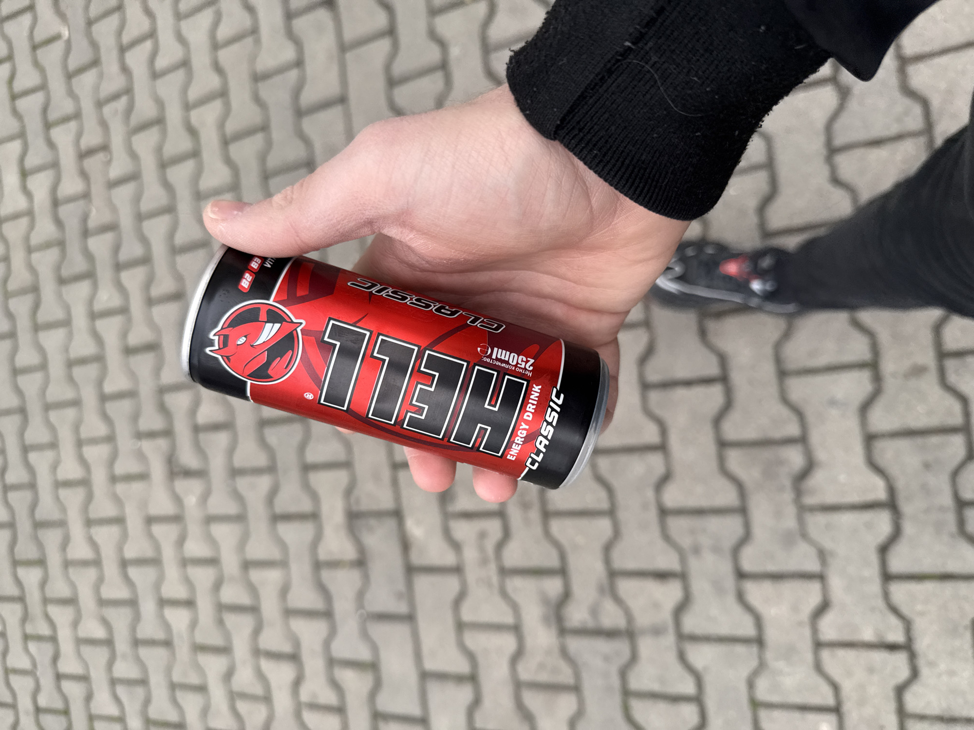 A person is holding a can of "HELL" energy drink. The can is red, black, and white, with the brand name prominently displayed. The person is wearing a black sweatshirt and black pants, with black shoes visible in the background. The person is standing on a paved surface made of gray bricks. The location is likely urban due to the paved surface and clothing, although no other definitive location clues are present. Stara Zagora, Bulgaria, cannot be confirmed as the location based on the information in the image.