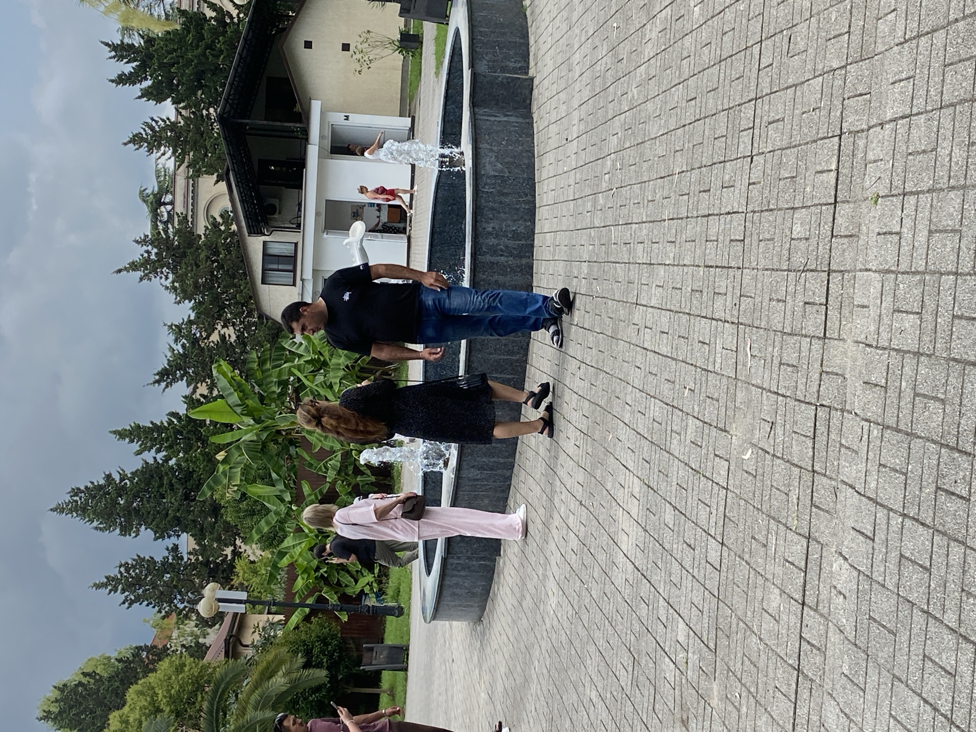 The image shows a paved area with a fountain in the background. Several people are visible, including a man in a black t-shirt and blue jeans standing in the foreground, and a woman in a pink outfit and another woman in a black dress standing near the fountain. Behind the fountain, there is a building with an open entrance. The environment appears to be outdoors with trees and vegetation. The sky is overcast. This scene is likely located in the city of Sokhumi, Abkhazia, given the context.