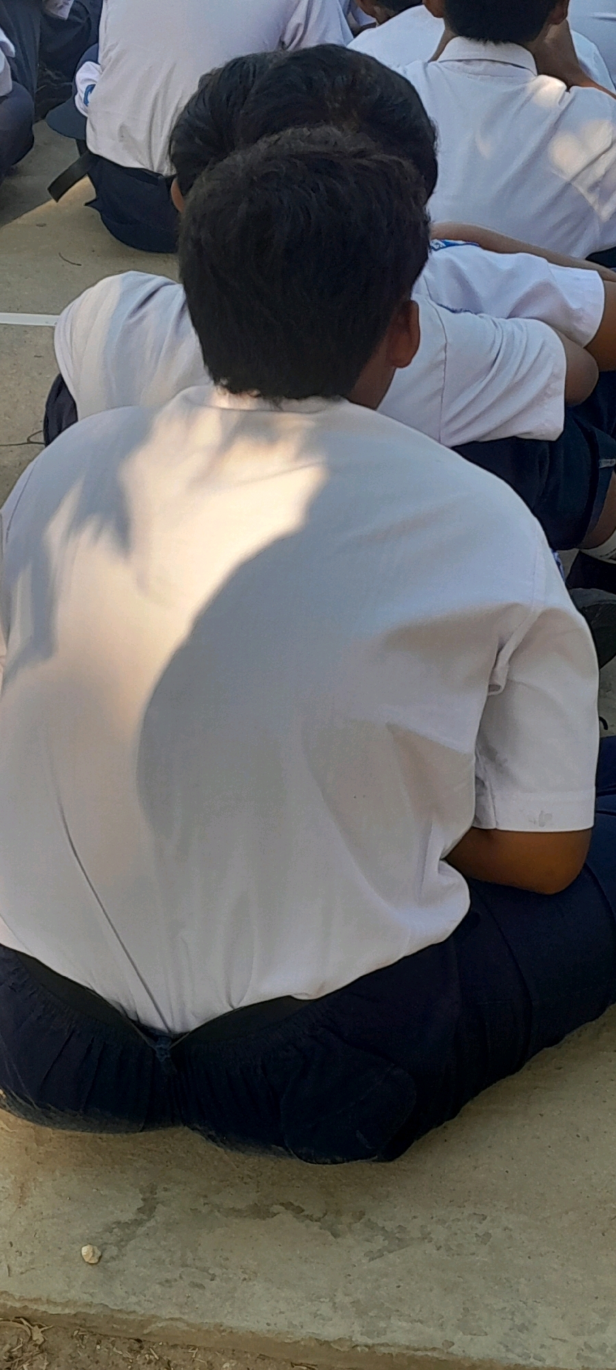 The image shows a close-up rear view of several people sitting on the ground, likely students based on their uniforms which consist of white shirts and dark blue pants or skirts. The focus is on the back of one individual in the foreground, who is wearing a white shirt and dark blue trousers. The person is sitting cross-legged on a textured, light-colored surface that appears to be concrete. Sunlight casts a shadow on the person's back, creating a dappled effect on their white shirt. In the background, other individuals in similar uniforms are visible, also sitting on the ground. The overall environment appears to be outdoors, possibly a schoolyard or an open public space. The location is identified as Tahunan, Indonesia.