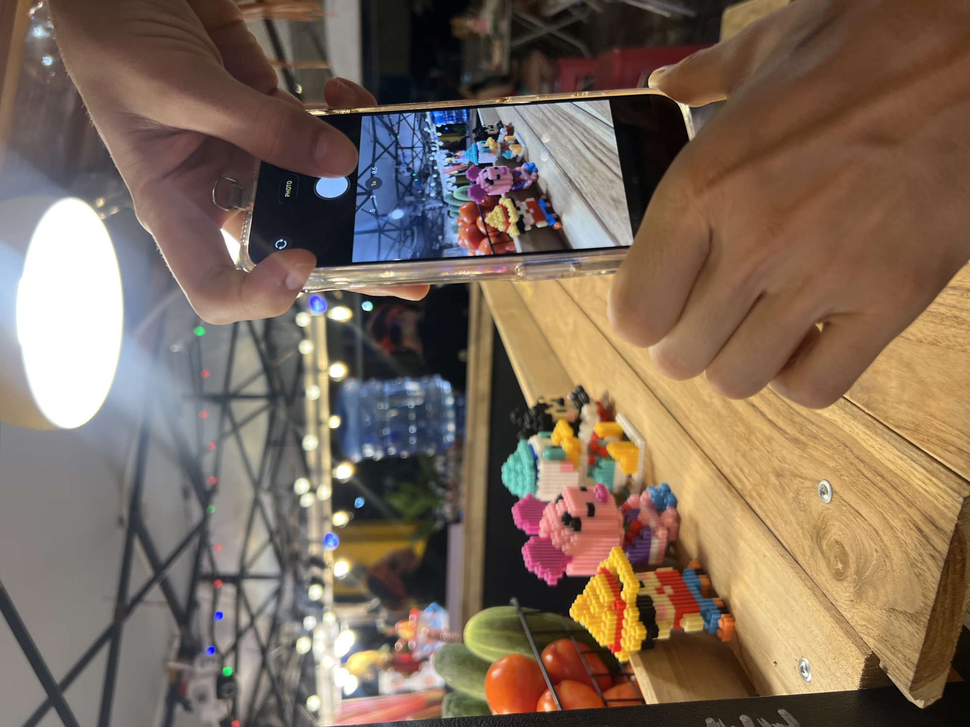 The media file shows a close-up of a person's hands holding a smartphone, capturing a photo of a collection of small, colorful, pixelated toy figures arranged on a wooden surface. The figures appear to be characters from various media, including what looks like Kirby, a cupcake, and other assorted small figurines. In the background, out of focus, are strings of lights, a large illuminated object (possibly a ring light), and what seems to be a market stall or food stand with produce like cucumbers and tomatoes visible. The scene is likely taking place in an indoor or covered outdoor setting, given the lighting and the market-like atmosphere. The time of day is unclear but the lighting suggests it could be evening or a dimly lit indoor space. The person's hands are positioned to take a photograph, indicating an activity of documenting or sharing. The image is a vertical orientation.