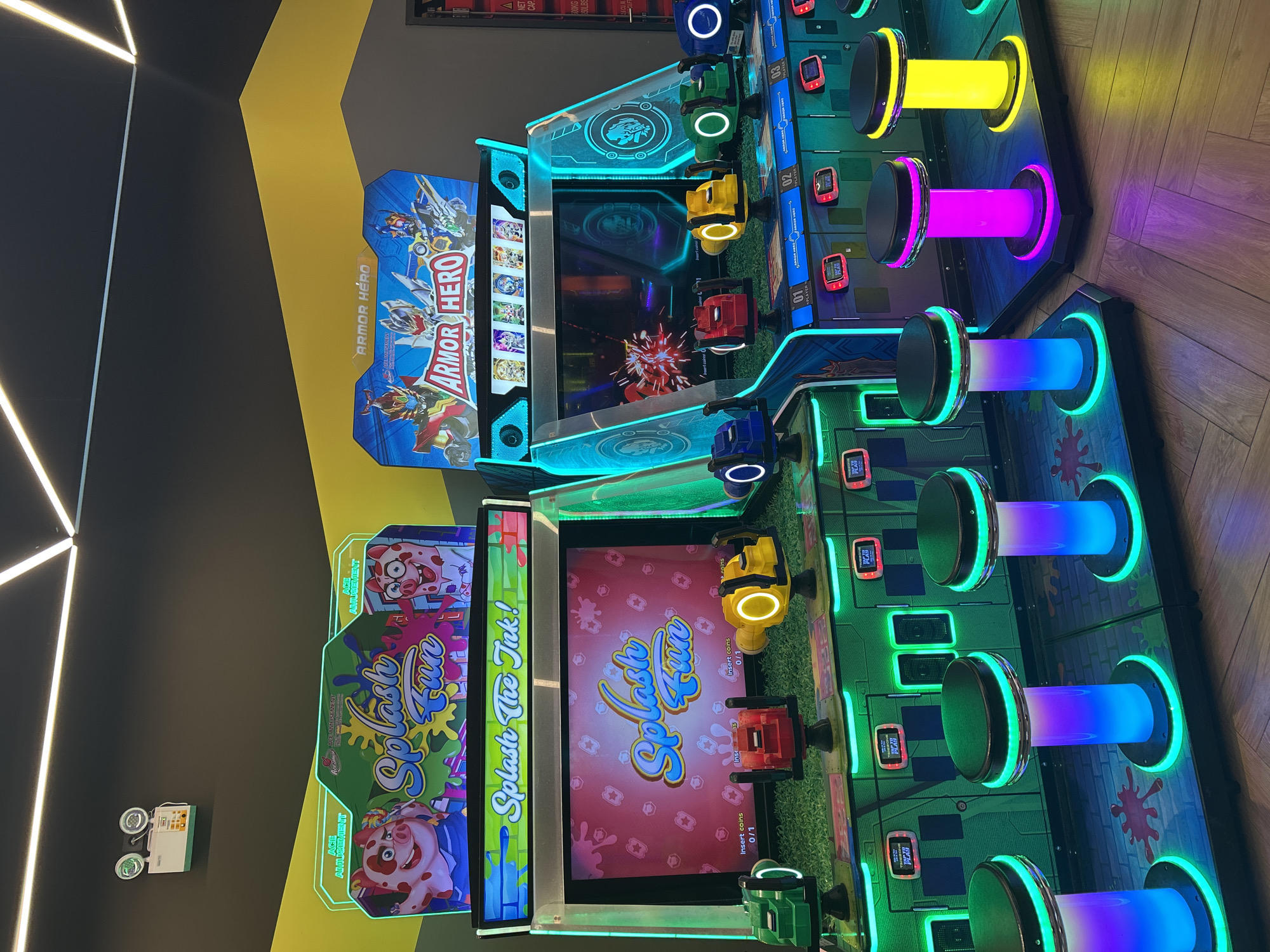 The image depicts a row of arcade game machines in an indoor entertainment venue. Two distinct games are visible: "Splash Fun" on the left, featuring cartoon pigs and water-based gameplay, and "Armor Hero" on the right, with images of armored characters. Each game station includes a large screen, input devices (likely water guns), and colorful, illuminated stools for players. The environment is dimly lit, with bright neon and LED lighting from the machines and ceiling fixtures creating a vibrant, modern atmosphere. The floor appears to be wooden parquet. The specific location within Makassar, Indonesia, is not provided in the image or its context.