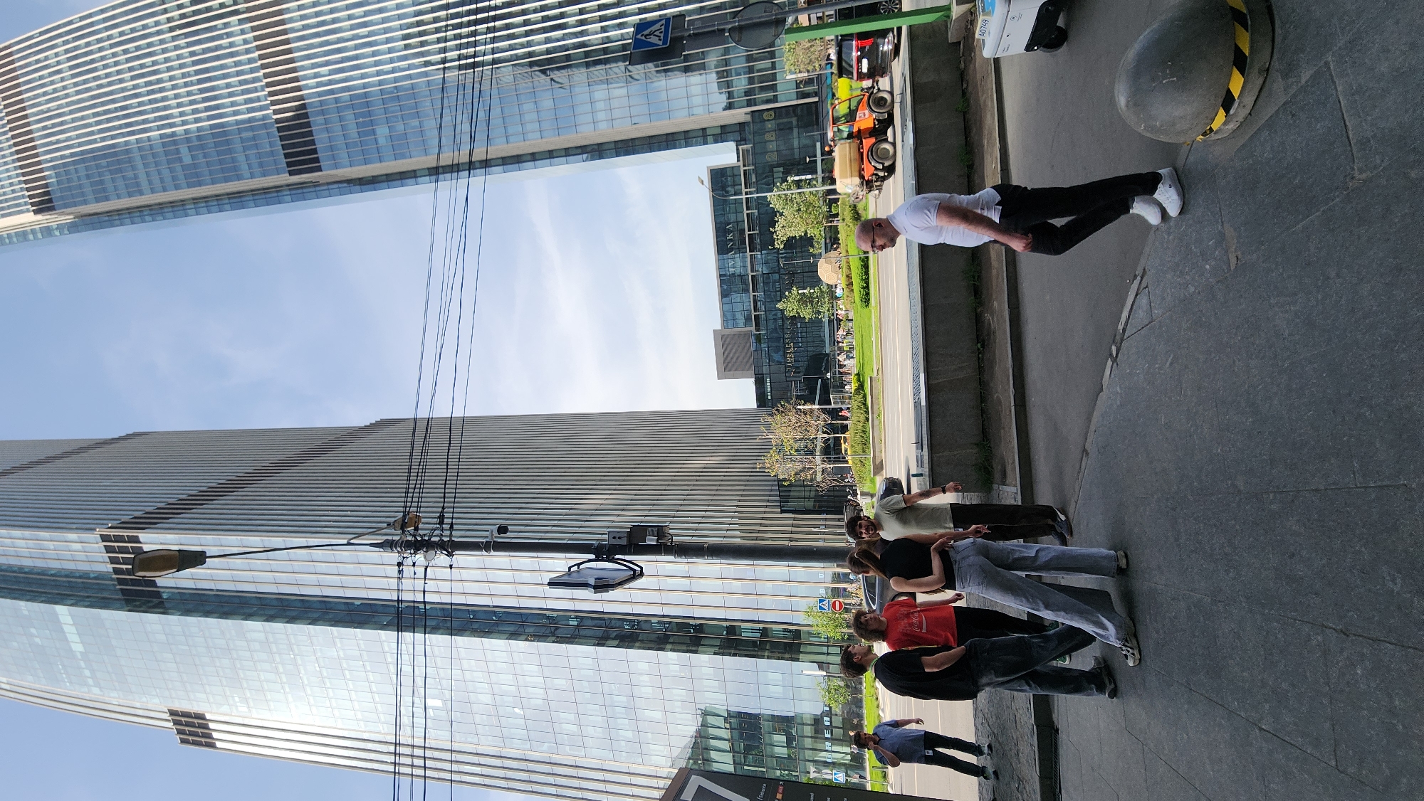 The image shows a street scene in Moscow, Russia, with a group of people standing on the sidewalk. A man in a white shirt and black pants is walking in the opposite direction. The scene is framed by modern glass skyscrapers. A street sign is visible overhead. There are electrical wires above the group. Several vehicles, including a small orange vehicle, are visible on the street. A concrete bollard with yellow and black stripes is on the sidewalk.