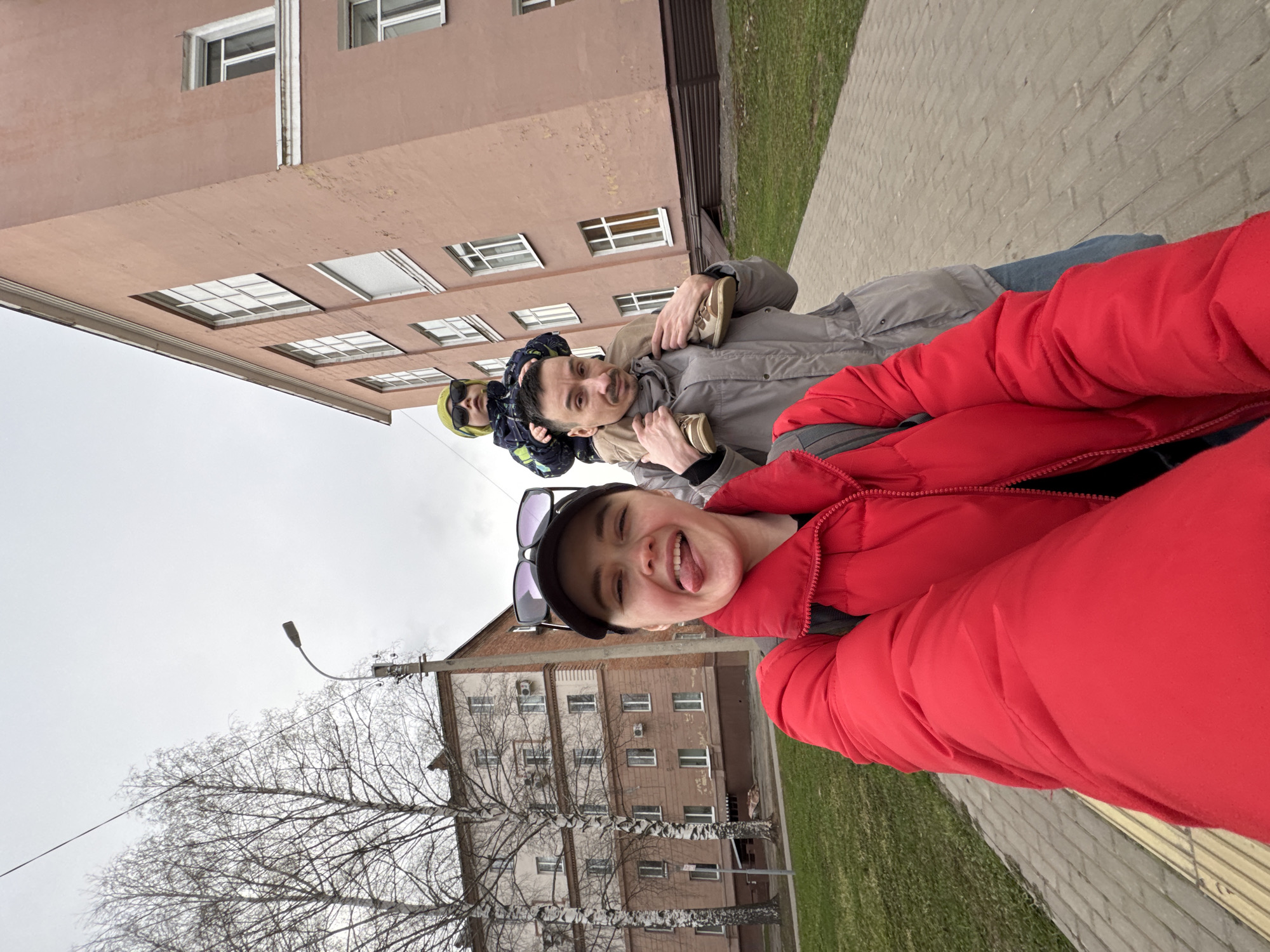 The image, oriented vertically, captures a playful outdoor moment featuring a group of people, likely a family or friends, in Izhevsk, Russia. In the immediate foreground, a person in a vibrant red puffer jacket and a black cap (with sunglasses resting on top) faces the camera, sticking out their tongue in a humorous gesture.
Behind them, a man in a grey jacket carries a young child on his shoulders; the child wears a yellow hat and sunglasses. Another person in a blue jacket is partially visible behind the man.
The background shows an urban environment with two multi-story buildings. One building is a light reddish-brown, featuring multiple white-framed windows, and the other, further back, is a lighter brown with similar window styles. The ground consists of a light grey paved sidewalk and a strip of green grass. Several bare trees with intricate branches suggest late winter or early spring. A streetlamp on a utility pole is also visible against the overcast sky, indicating daytime with diffused light. The heavy outerwear worn by the individuals confirms cooler weather conditions. No visible text is present in the image.