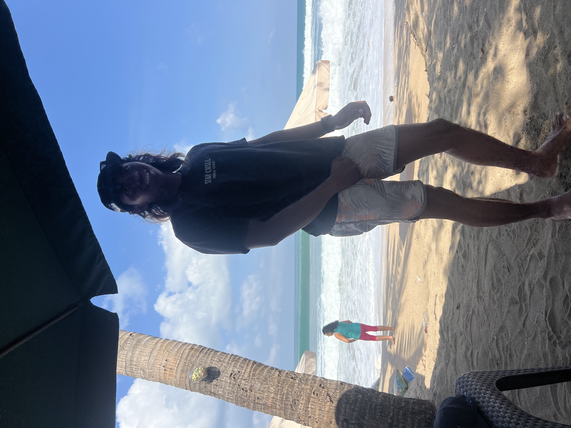 An outdoor, eye-level, medium shot captures a young man standing on a sandy beach in Kuta, Indonesia. He is wearing a dark t-shirt with white lettering that reads "STAY CHILL" and patterned shorts. The man is facing forward with his left hand near his groin and his right arm is bent at the elbow. He is looking at the camera and smiling. His hair is long and dark, and he is wearing a dark baseball cap.
Behind him, the ocean stretches out under a bright blue sky with scattered white clouds. Gentle waves are breaking on the shore. A palm tree is visible to the left of the man, and a beach umbrella casts shadows on the sand. In the background, another person can be seen standing near the water's edge, wearing a teal tank top and pink leggings. Beach chairs and a water bottle are scattered on the sand.
The lighting is bright, suggesting it is daytime. The overall atmosphere is relaxed and tropical.