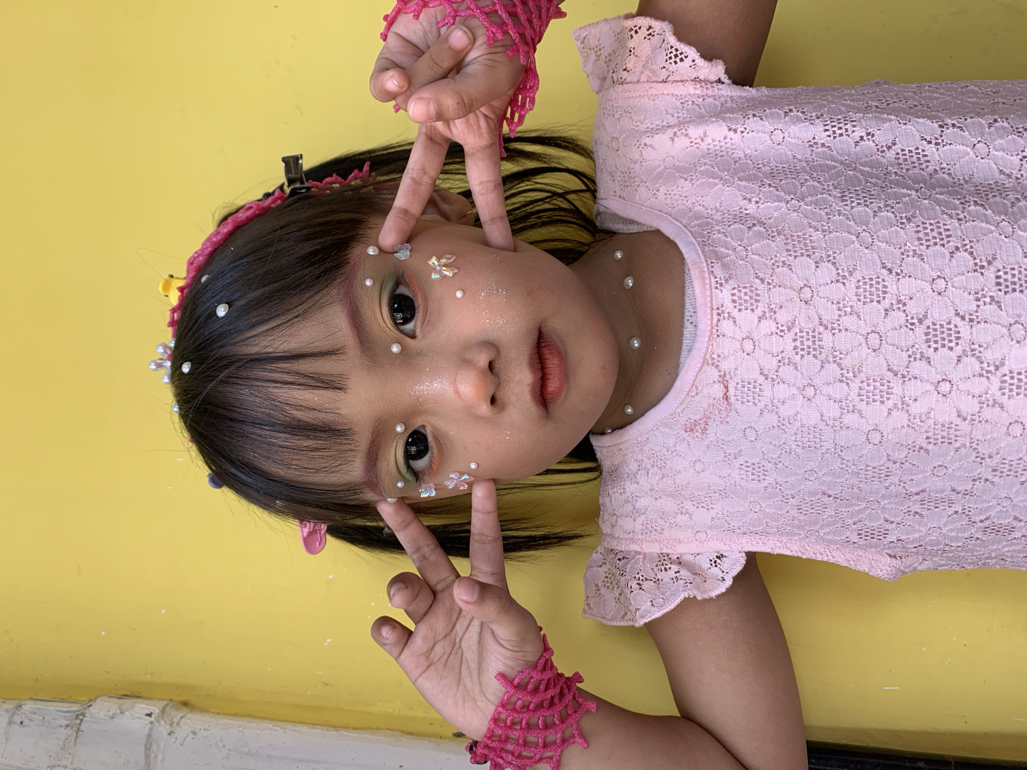 A young girl, likely of Southeast Asian descent, is pictured posing against a bright yellow wall. She has dark hair styled into two pigtails and is adorned with small pearls and glitter on her face, giving her a whimsical appearance. She is wearing a pink lace dress with ruffled sleeves and a white undershirt. Her right hand is held up near her face, with her index and middle fingers extended in a peace sign, while her left hand is also raised, fingers spread. On her wrists, she wears bright pink, net-like bracelets. The background is a simple, textured yellow wall, suggesting an outdoor setting, potentially in an urban environment like Malang, Indonesia, where similar vibrant walls are common in residential or commercial areas. The lighting appears to be natural daylight, casting soft shadows.