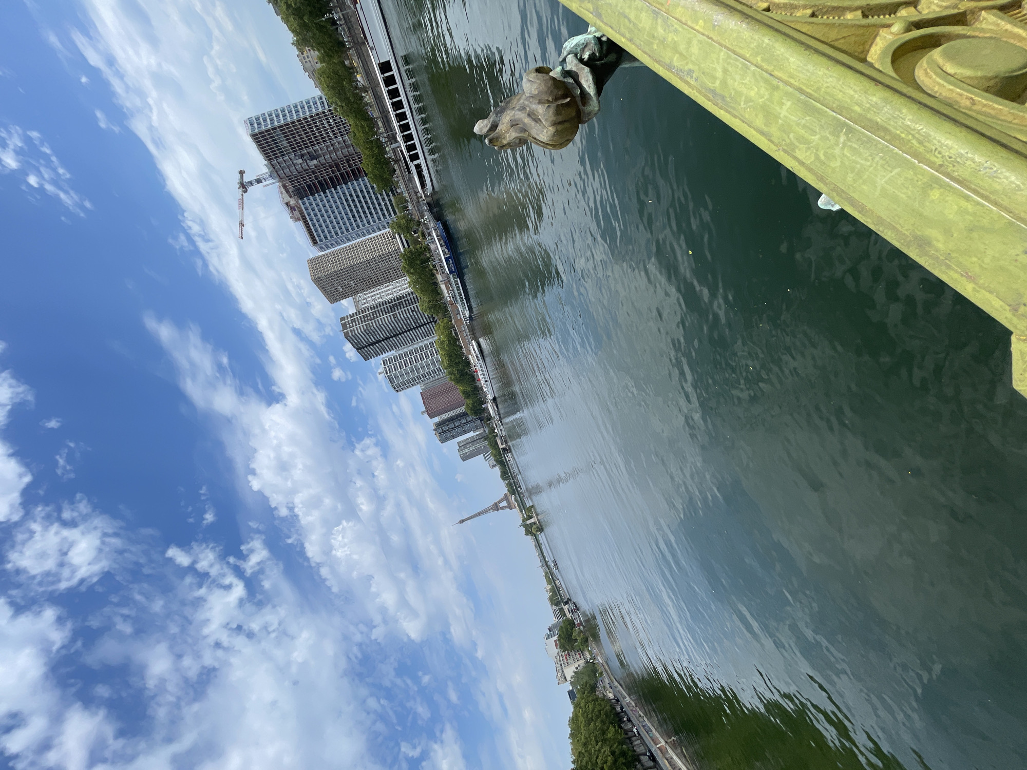 A wide-angle, low-angle shot captures a view across the Seine River in Paris, France, under a partly cloudy blue sky. In the foreground, a green, ornate bridge railing with graffiti is visible on the right. A bronze sculpture of a hand holding a flower adorns the railing on the far right. The river occupies the majority of the frame, its surface rippled and reflecting the sky and surrounding buildings.
On the far bank of the river, a line of trees runs along the edge, followed by a row of modern, tall buildings. One building in the distance is under construction, with a prominent crane visible at its peak. Further down the river, the iconic Eiffel Tower stands tall. Several boats, including tour boats and what appear to be houseboats, are docked or cruising on the river. The overall scene depicts a bright, urban day in Paris with a mix of historical landmarks and modern development.