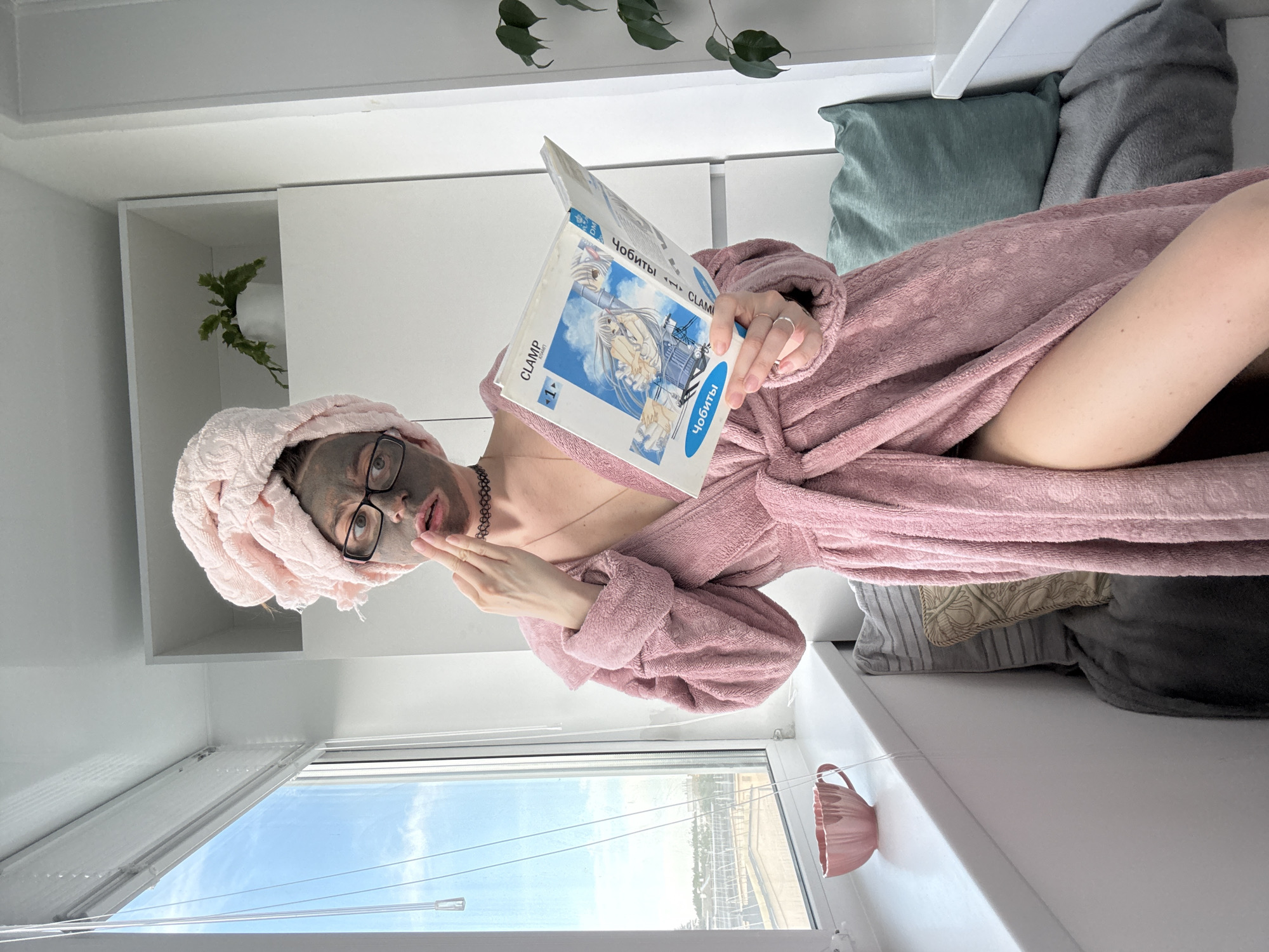 In an indoor setting near a window, a young woman with a pink towel turban and a face mask is reading a book. She is wearing a pink bathrobe and a black choker necklace. The room appears to be a balcony or a room with a large window, featuring a plant on a shelf and a pink teacup on the windowsill. The woman's gesture suggests she is interacting with the face mask. The overall lighting is bright, indicating daylight.