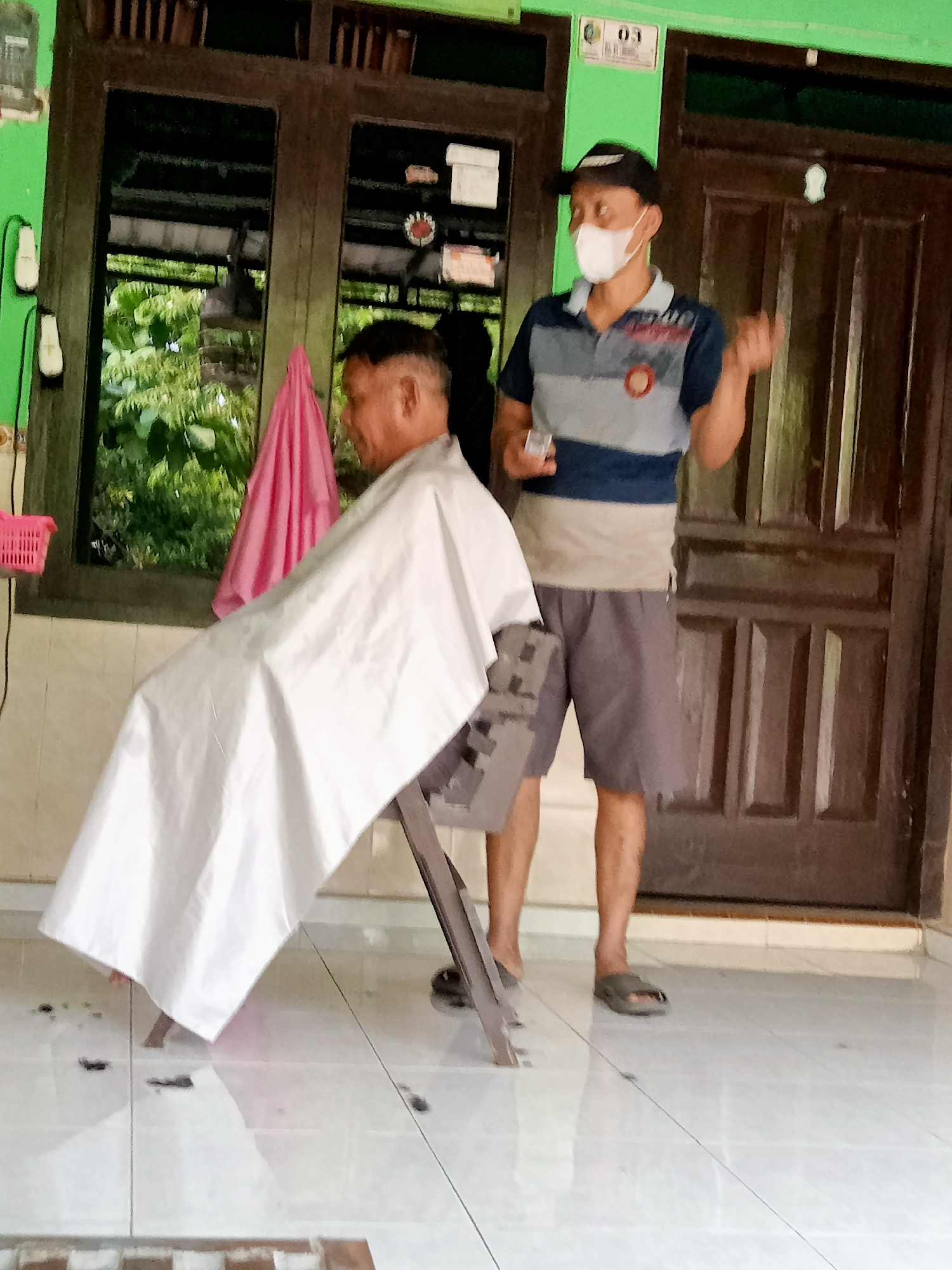 Two men are in front of a wooden door, possibly in a residential area of Majan, Indonesia. The man on the left, wearing a white cape, is seated and appears to be receiving a haircut. His hair is short and styled in an undercut. The man on the right, standing and wearing a baseball cap and a face mask, is holding a small object, likely a pair of scissors, and gesturing with his other hand. He is dressed in a striped polo shirt and shorts. The environment appears to be an outdoor porch or covered walkway, with tiled flooring and glimpses of greenery and a pink fabric hanging in the background.
