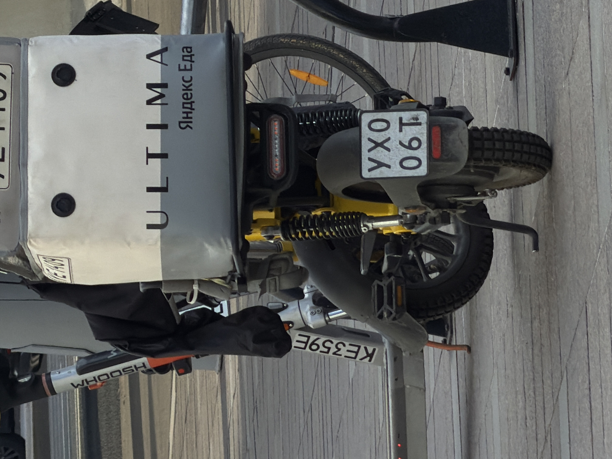 The image captures a close-up, slightly angled view of a parked electric bicycle in an urban setting. The bike features a prominent delivery box labeled "ULTIMA Яндекс Еда" (Yandex Food) on its rear carrier. The license plate, partially obscured, reads "YX0 06T." Another license plate, "KE359E," is visible below the frame. The bike is branded "WHOOSH" on its frame. The backdrop shows a textured wall or facade, suggesting an urban environment. The overall scene depicts a delivery service vehicle in an undefined location.