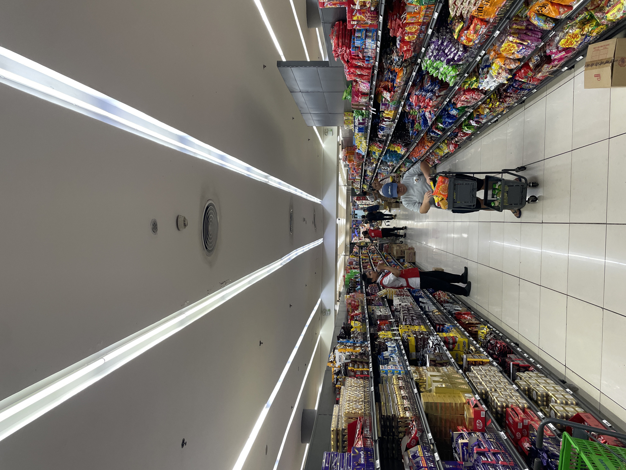 A full shot of a well-lit supermarket aisle, likely in Davao City, Philippines, features a wide variety of consumer goods displayed on tall shelving units that extend into the distance. The ceiling is fitted with long, bright fluorescent light fixtures.
In the foreground, a person wearing a blue cap and a grey t-shirt pushes a shopping cart filled with groceries. The cart itself is a dark grey color with two levels for storage.
Further down the aisle, a supermarket employee, dressed in a red vest over a white shirt and black pants, is seen attending to the shelves. Other shoppers are visible in the background, some browsing products and others walking through the aisle. The floor is tiled in a light color. The shelves are stocked with numerous colorful packages of snacks and confectionery.